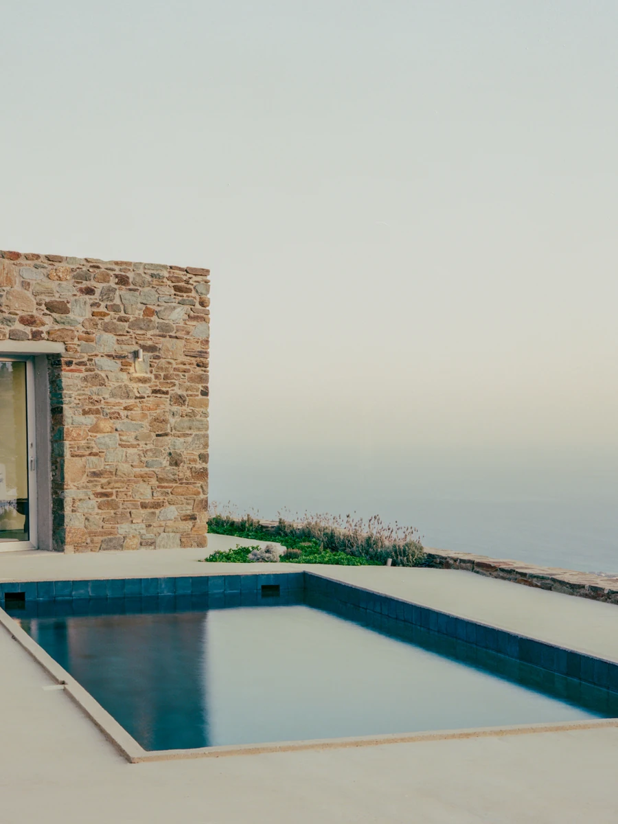 Tinos House by Kelly Spanou, photographed by Lorenzo Zandri — A rectangular lap pool set into a pale concrete terrace, with a schist stone volume and full-height sliding glass to the left and the sea filling the horizon.