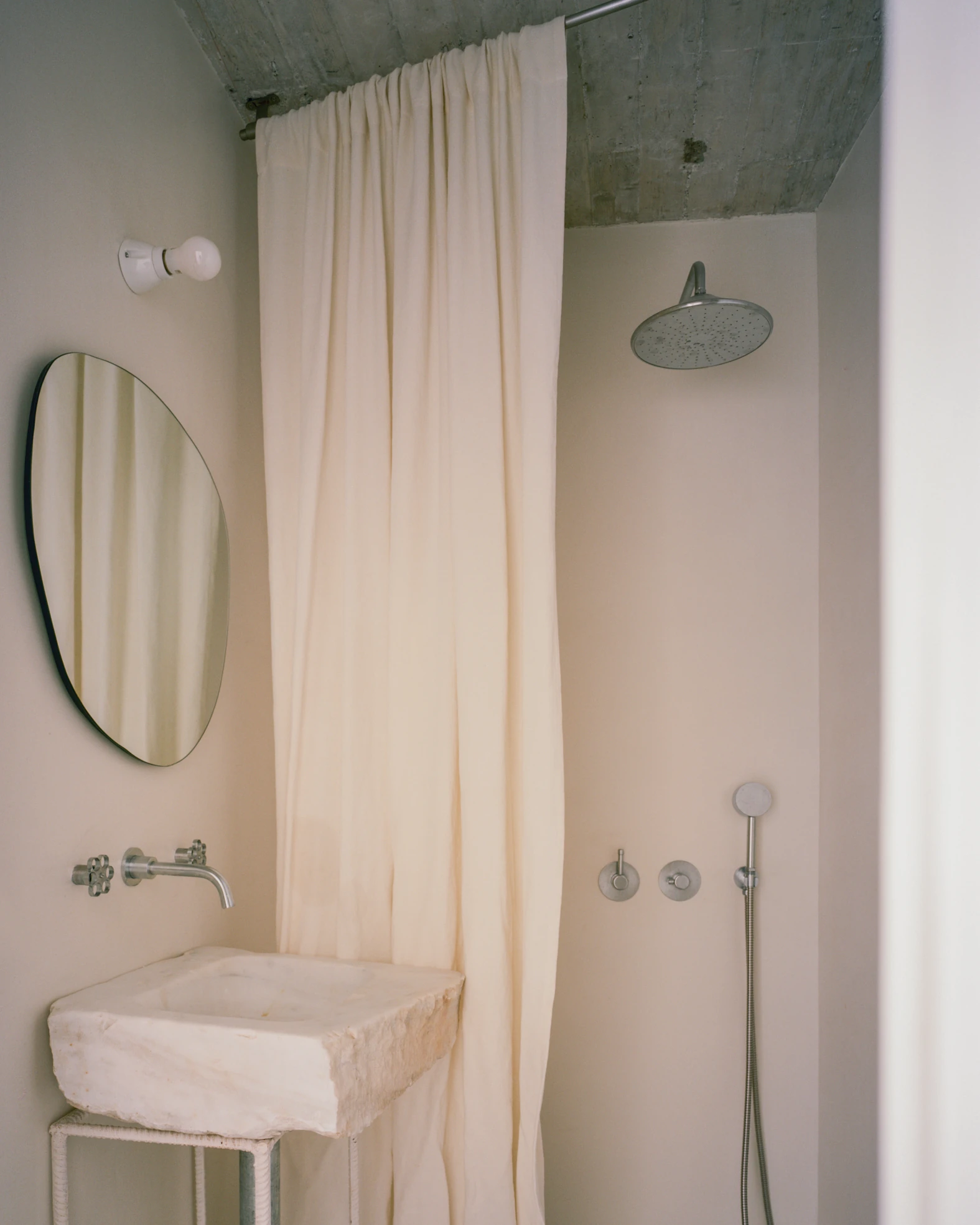 Tinos House by Kelly Spanou, photographed by Lorenzo Zandri — A reclaimed marble block sink with a rough hewn edge, wall-mounted steel taps, an organic-shaped mirror and a linen curtain drawn beside the shower.