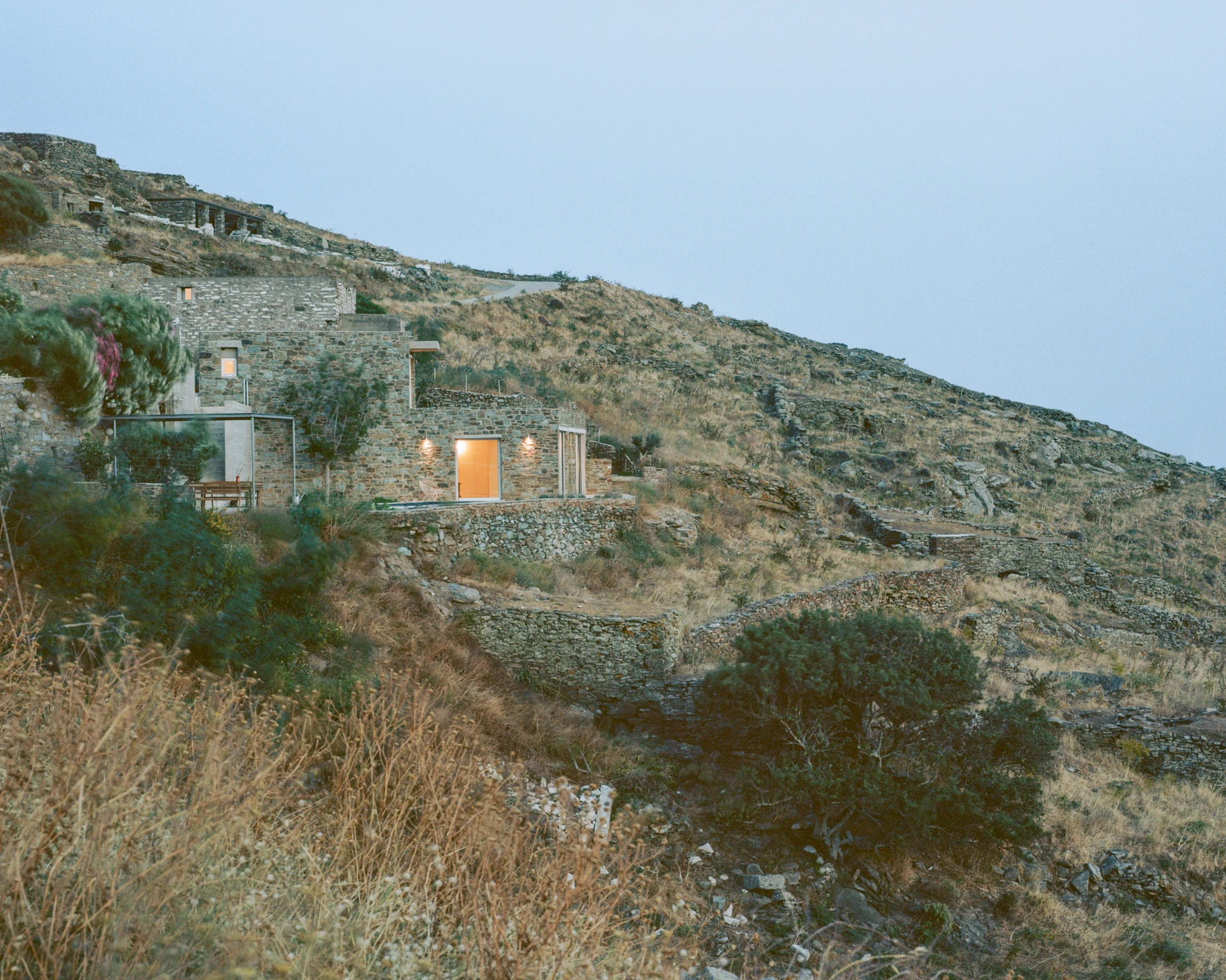 Tinos House by Kelly Spanou, photographed by Lorenzo Zandri — The house at dusk on a terraced hillside, its stone volumes stepping down between dry stone retaining walls, one window lit orange.