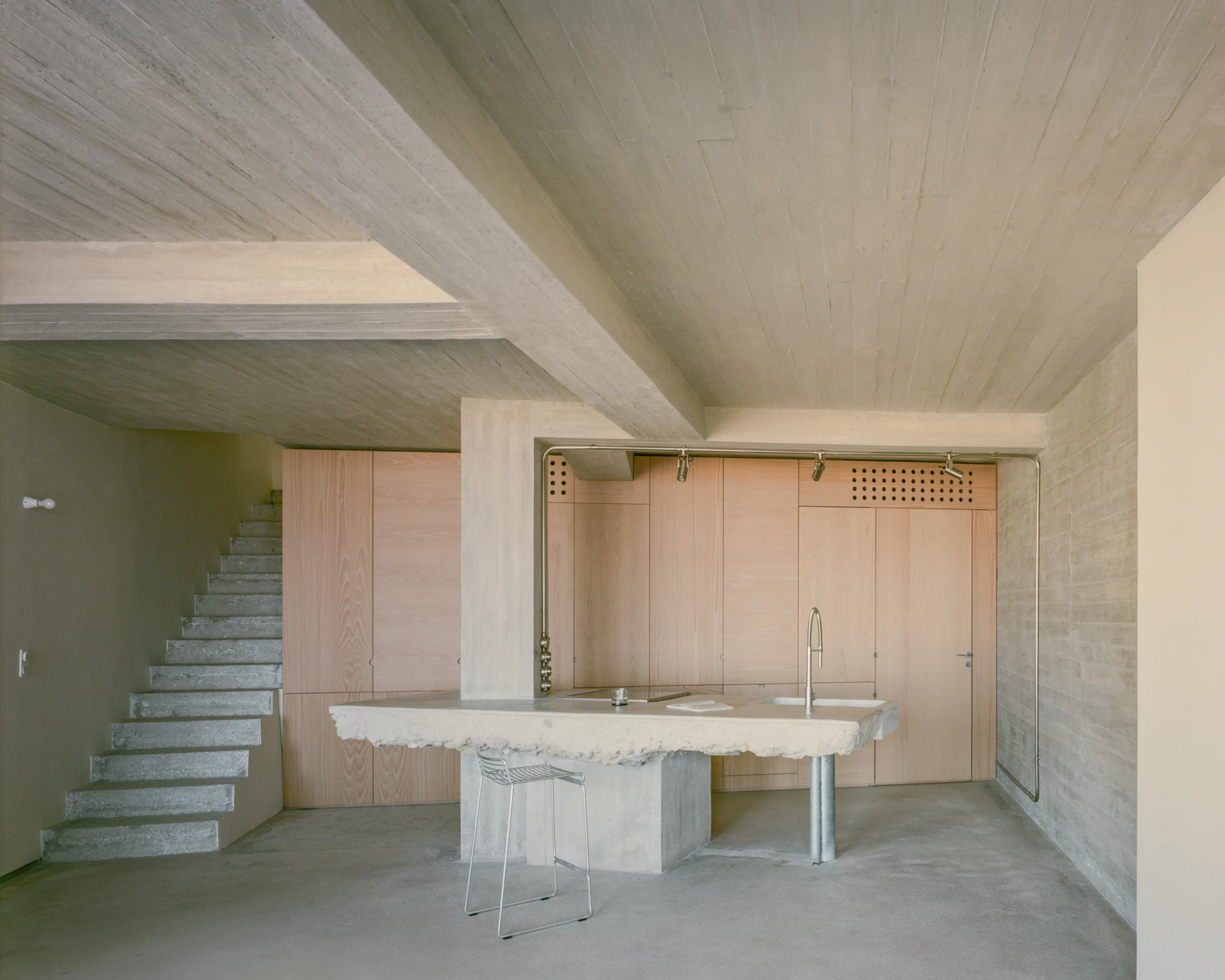 Tinos House by Kelly Spanou, photographed by Lorenzo Zandri — The ground floor monolith: a thick marble slab counter with a fractured raw edge on a concrete base, pale ash cabinetry behind and a cantilevered concrete stair to the left.