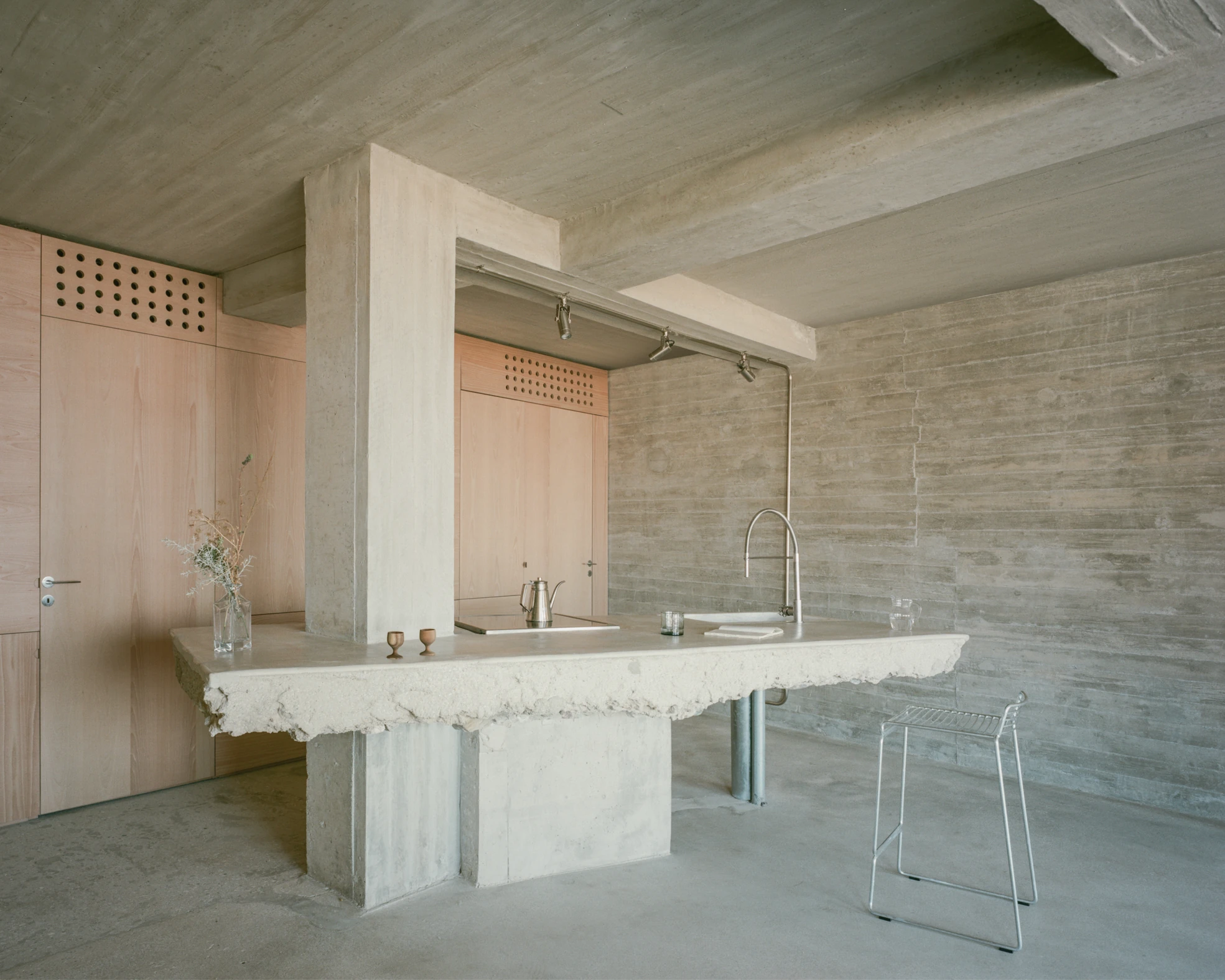 Tinos House by Kelly Spanou, photographed by Lorenzo Zandri — A concrete column passes through the marble counter of the kitchen monolith, with steel track lighting overhead and board-formed concrete walls behind.