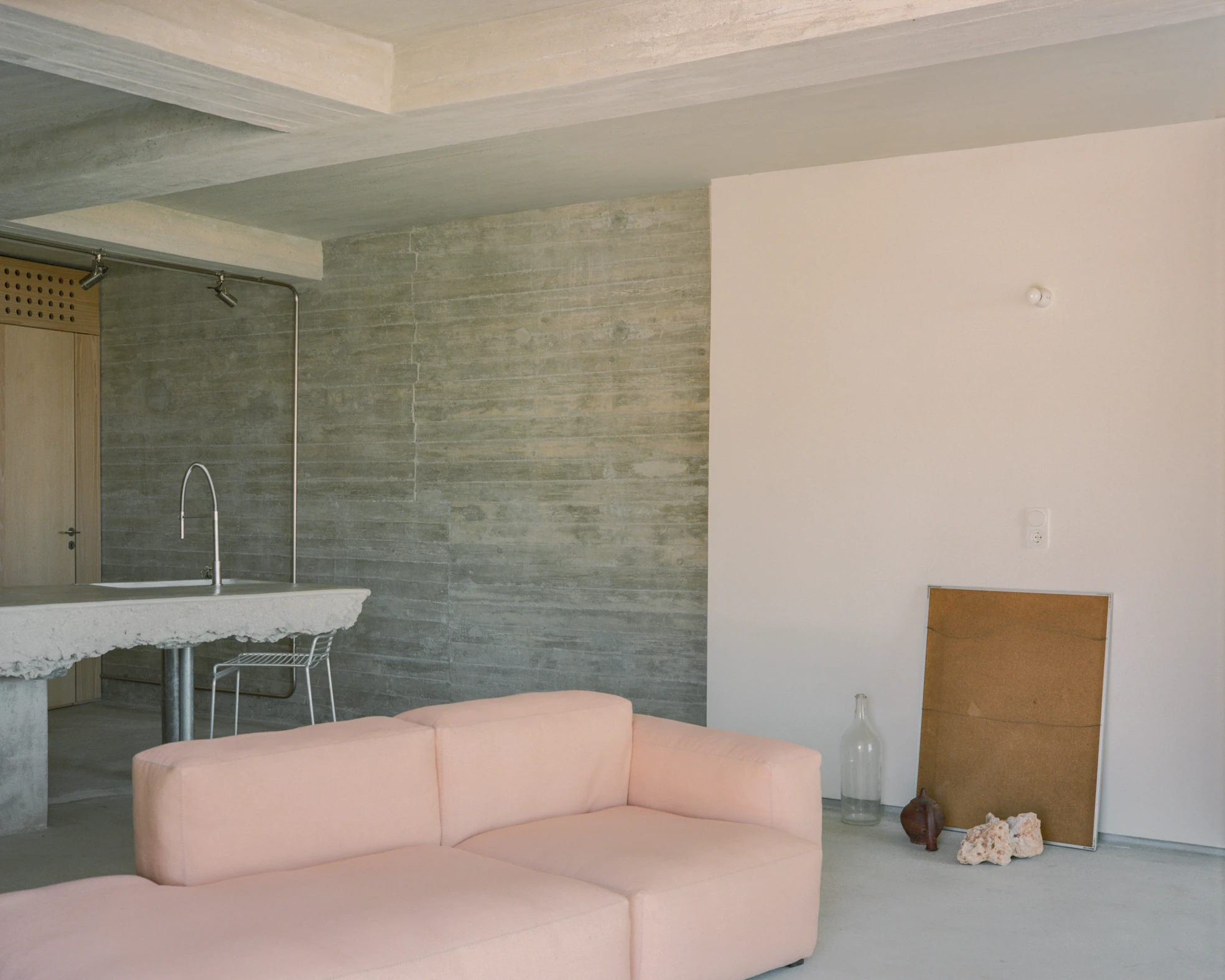 Tinos House by Kelly Spanou, photographed by Lorenzo Zandri — A pale pink modular sofa in front of a board-formed concrete wall, with the marble counter and its raw broken edge visible through the opening beyond.