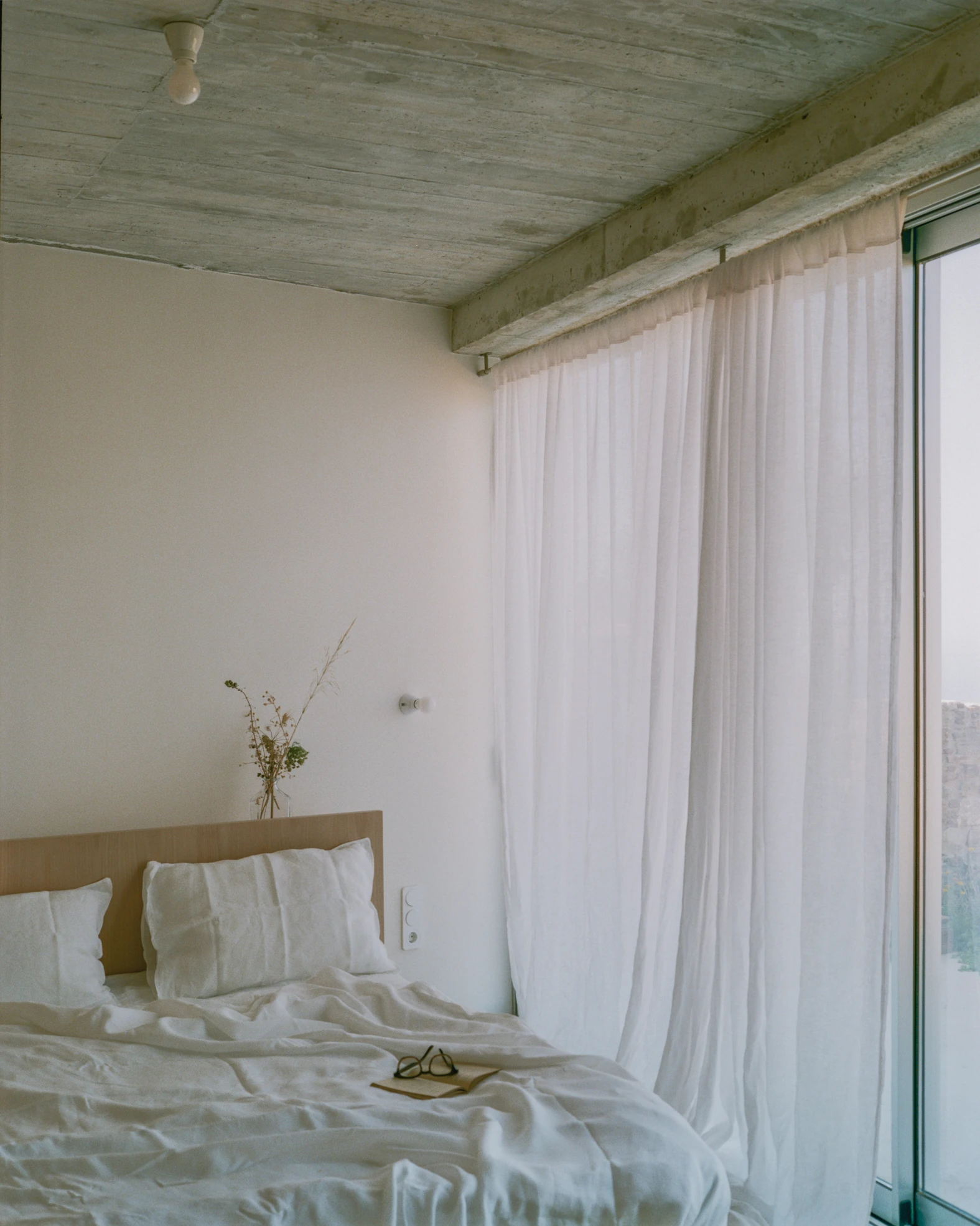 Tinos House by Kelly Spanou, photographed by Lorenzo Zandri — A bed with white linen below a board-formed concrete slab ceiling, sheer curtains hung the full height of the glazed wall.