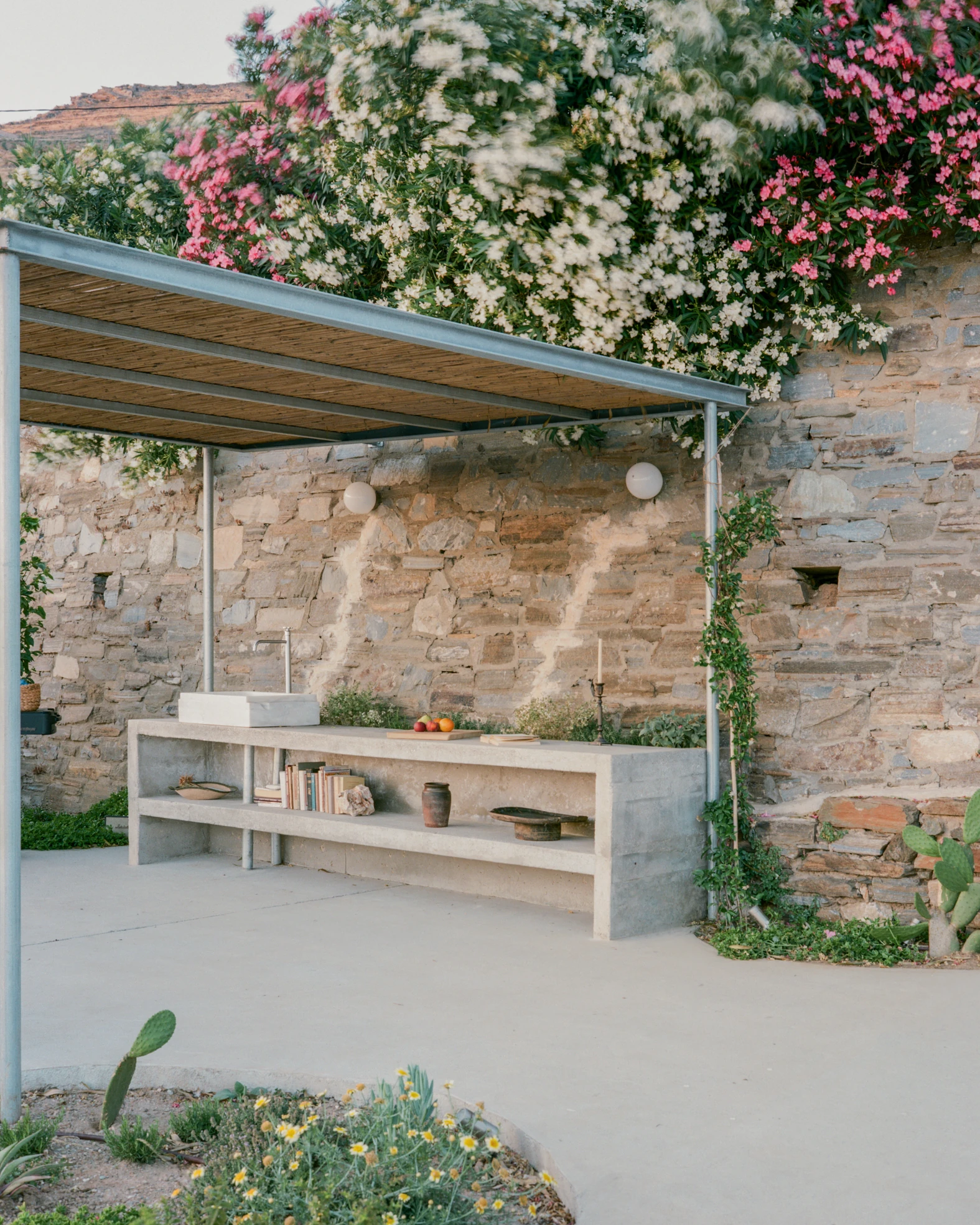 Tinos House by Kelly Spanou, photographed by Lorenzo Zandri — An outdoor kitchen under a reed and steel canopy, with a concrete counter and stone sink against a rubble wall, oleander in flower above.