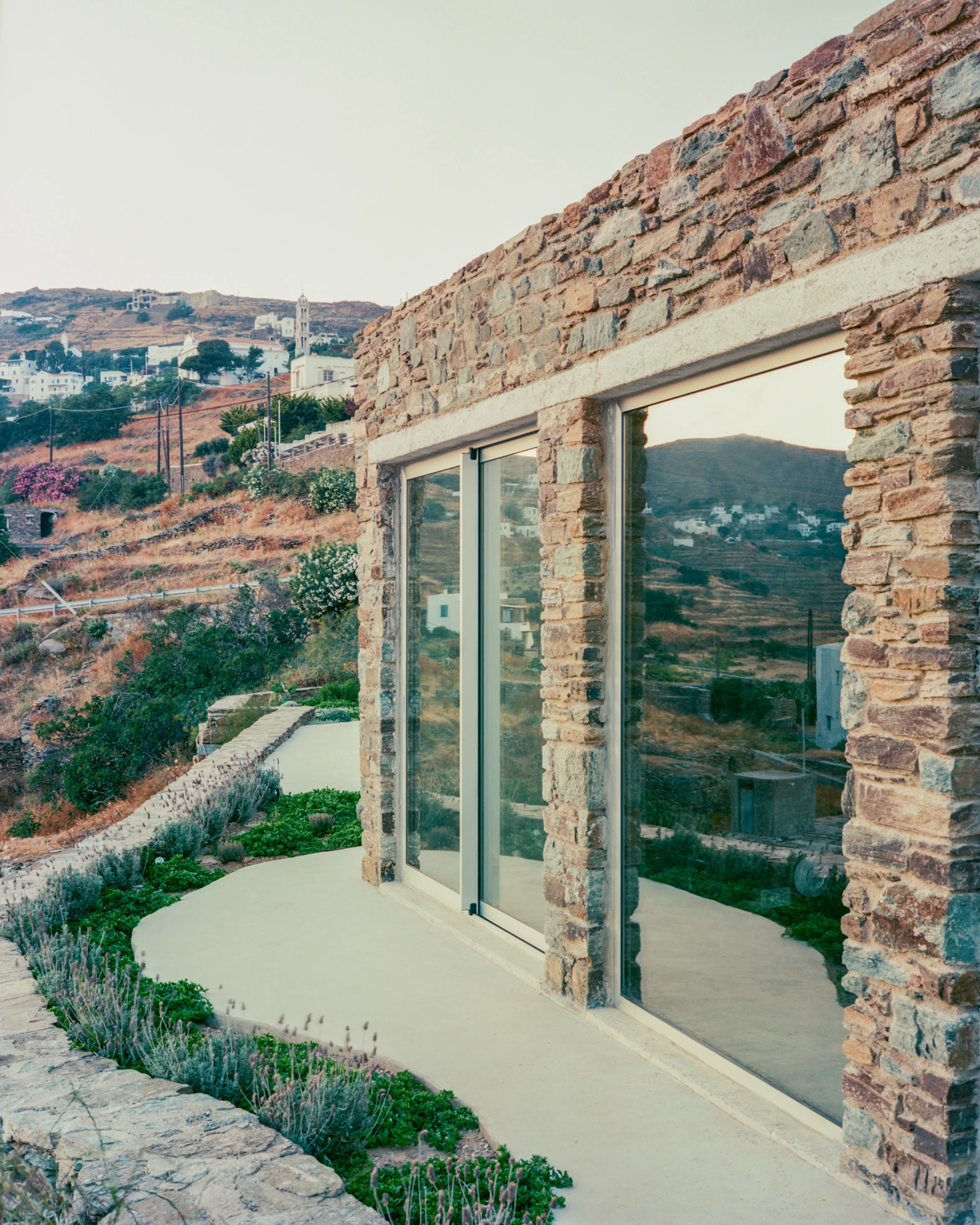 Tinos House by Kelly Spanou, photographed by Lorenzo Zandri — A full-height sliding glass panel set into a schist wall, reflecting the terraced hillside and the whitewashed village opposite.