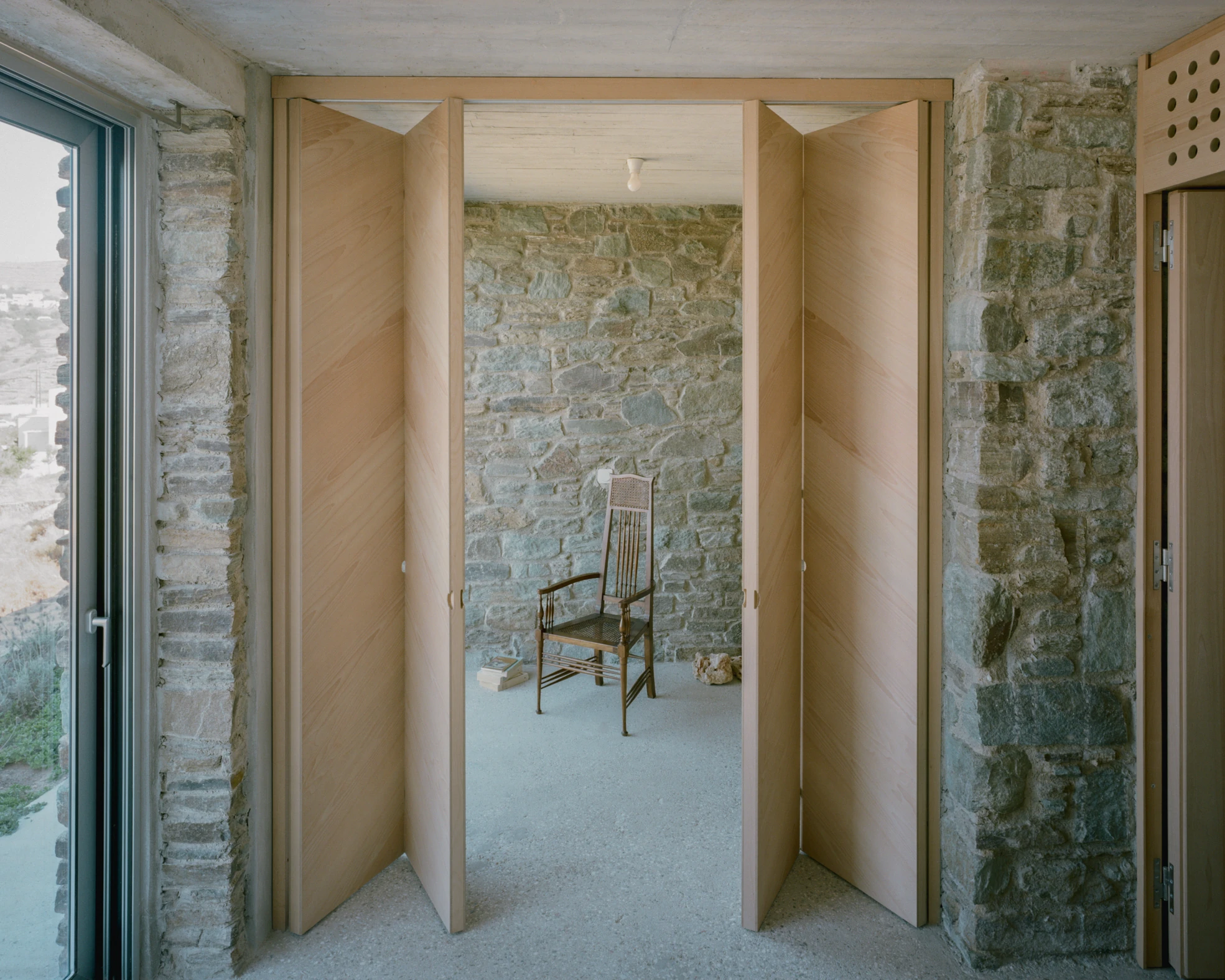 Tinos House by Kelly Spanou, photographed by Lorenzo Zandri — Folding ash doors opened onto a room with an exposed schist wall, a caned wooden chair and a stack of books on the concrete floor.