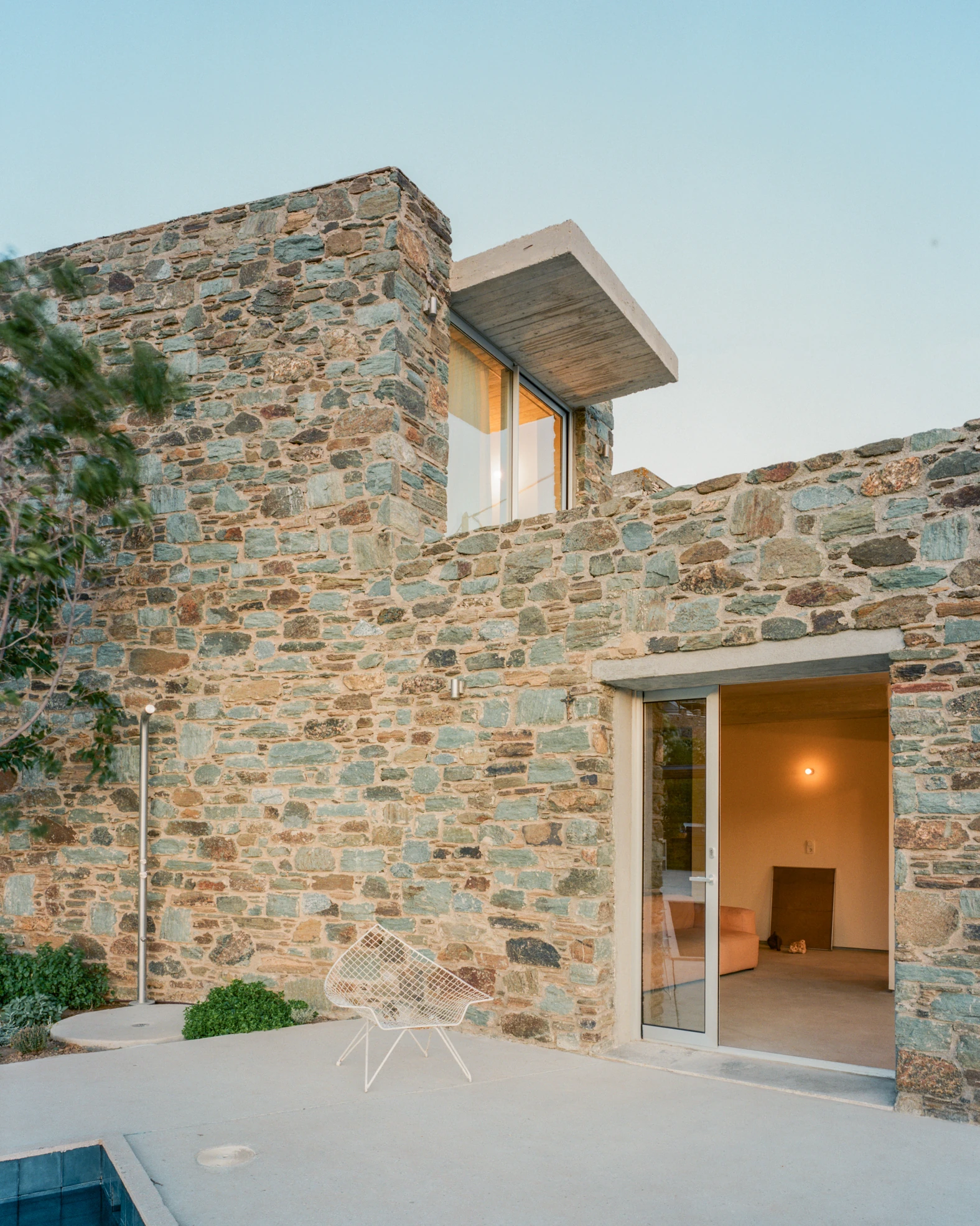 Tinos House by Kelly Spanou, photographed by Lorenzo Zandri — A two-storey schist facade with a cantilevered concrete canopy over an upper window, a white wire chair on the terrace and an open sliding door showing a lit room.