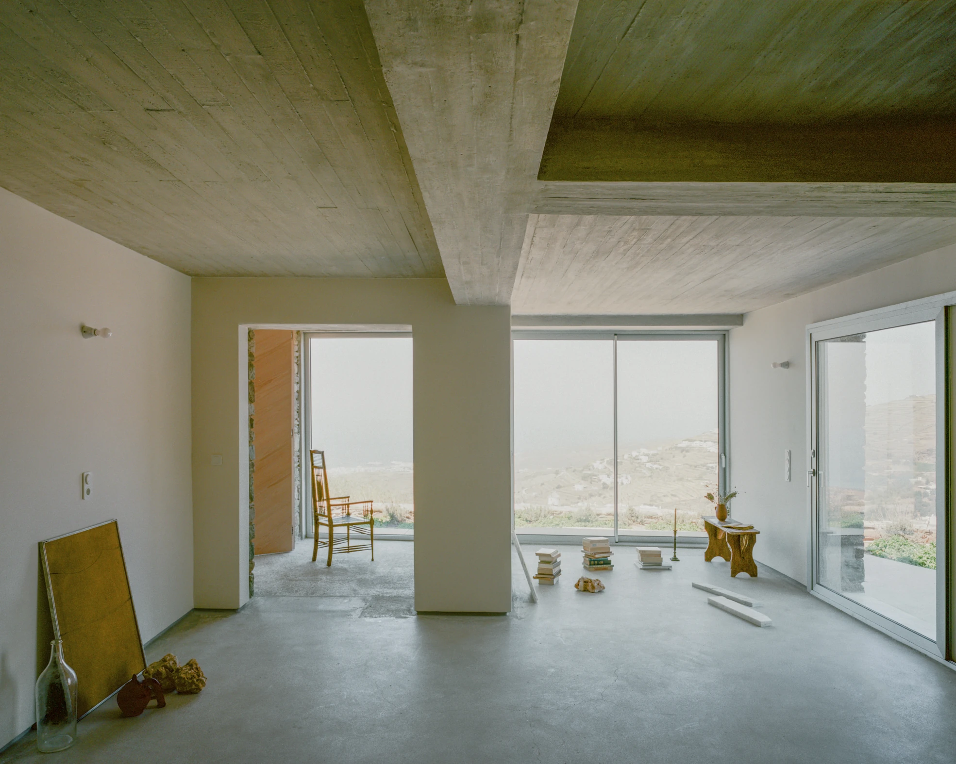 Tinos House by Kelly Spanou, photographed by Lorenzo Zandri — A room glazed on two sides, looking out to terraced fields and a whitewashed village, with books stacked on the concrete floor and a small wooden bench.