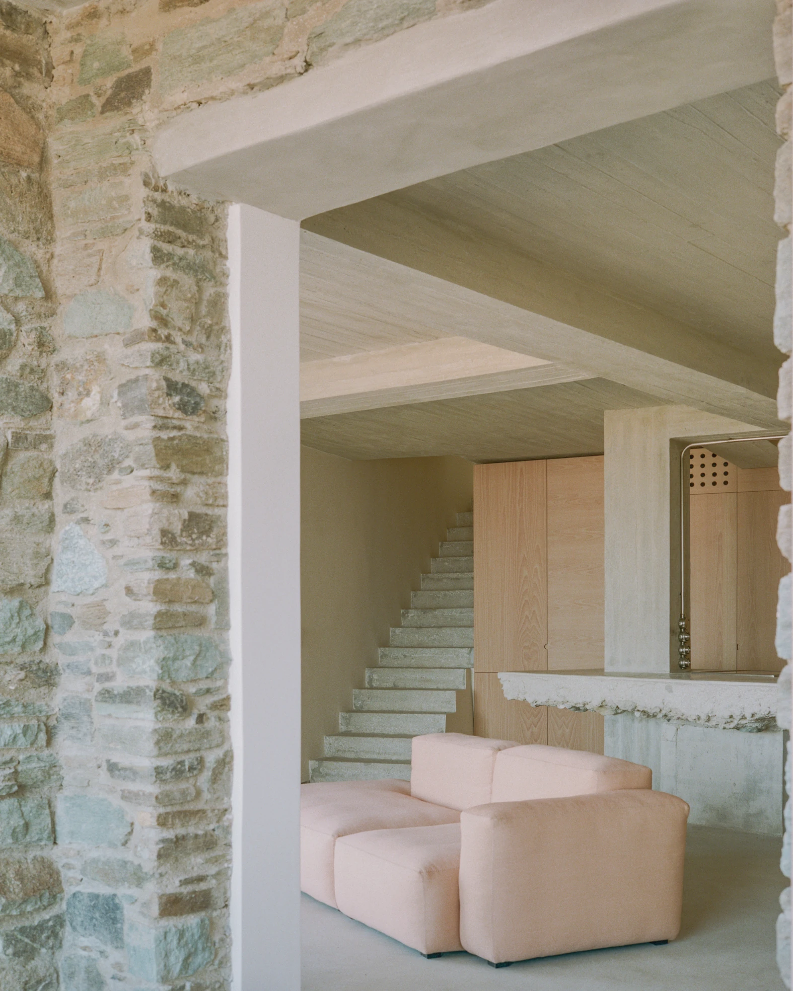 Tinos House by Kelly Spanou, photographed by Lorenzo Zandri — Looking through a schist opening into the living room, where a cantilevered concrete stair rises beside ash cabinetry and a pale pink modular sofa.