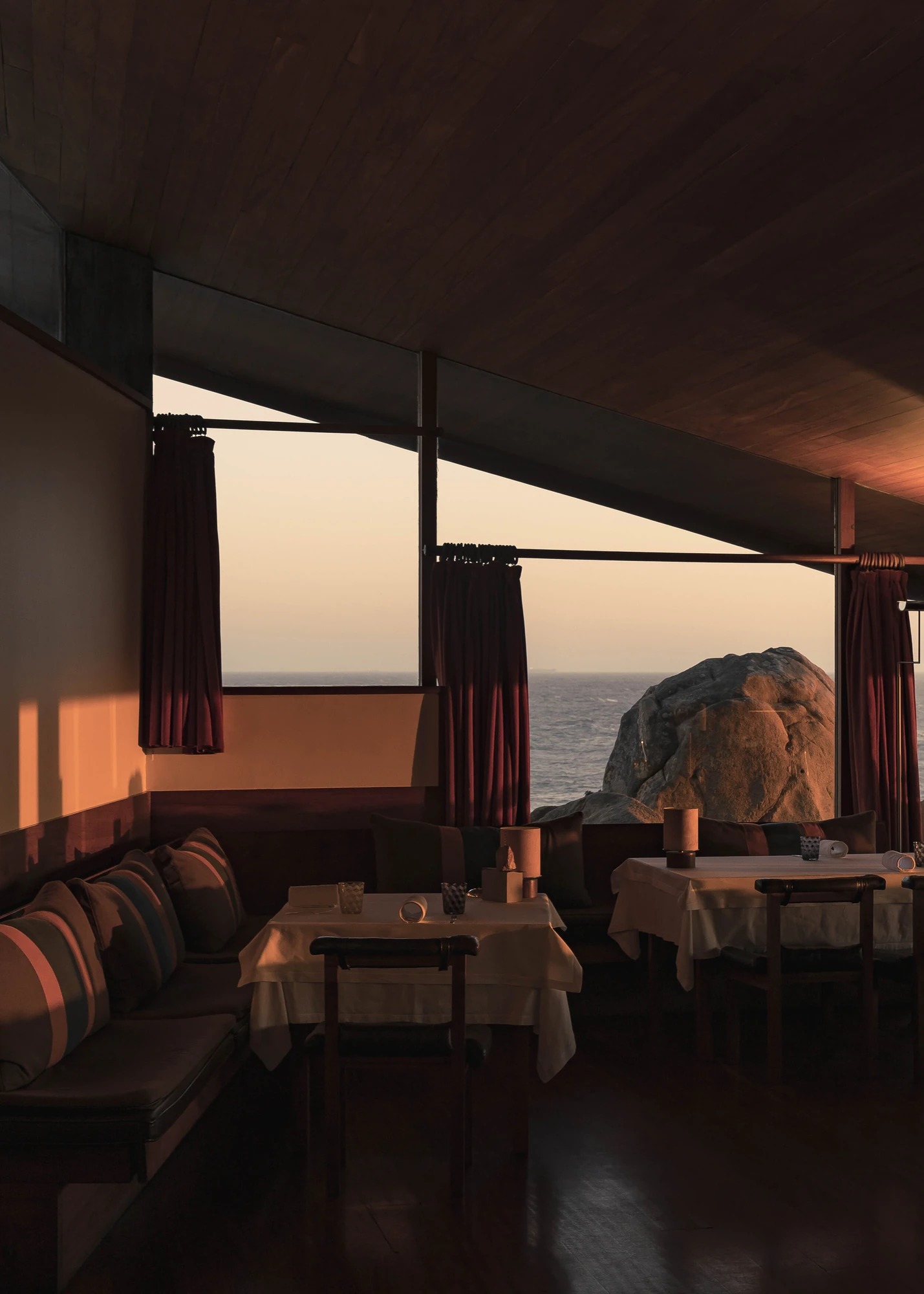 Casa de Chá da Boa Nova by Álvaro Siza, photographed by Nick Dearden — The dining room at sunset, with a built-in banquette and striped cushions, tables in white cloth, wine-red curtains and a granite boulder filling the window.