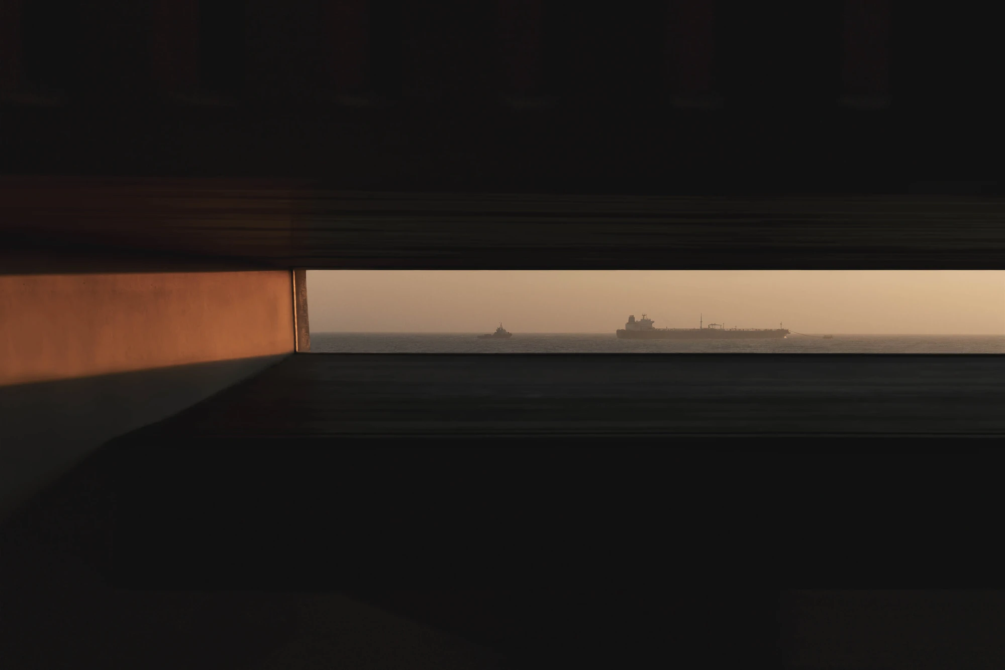 Casa de Chá da Boa Nova by Álvaro Siza, photographed by Nick Dearden — A narrow horizontal slot of window in a dark interior, framing a tanker and a small tug on a hazy horizon, with warm light raking one rendered wall.