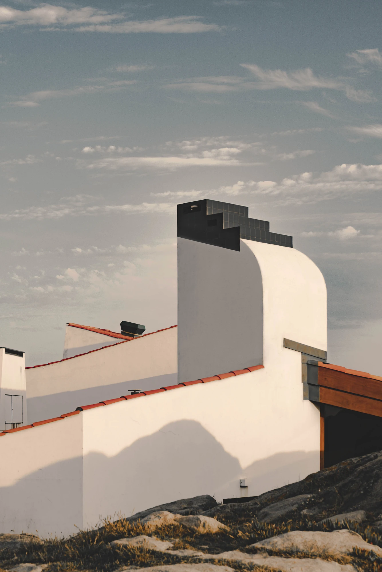 Casa de Chá da Boa Nova by Álvaro Siza, photographed by Nick Dearden — Roofscape of white rendered walls and red ridge tiles, with a black tiled chimney stack stepping against the sky above a wall rolled into a curve.