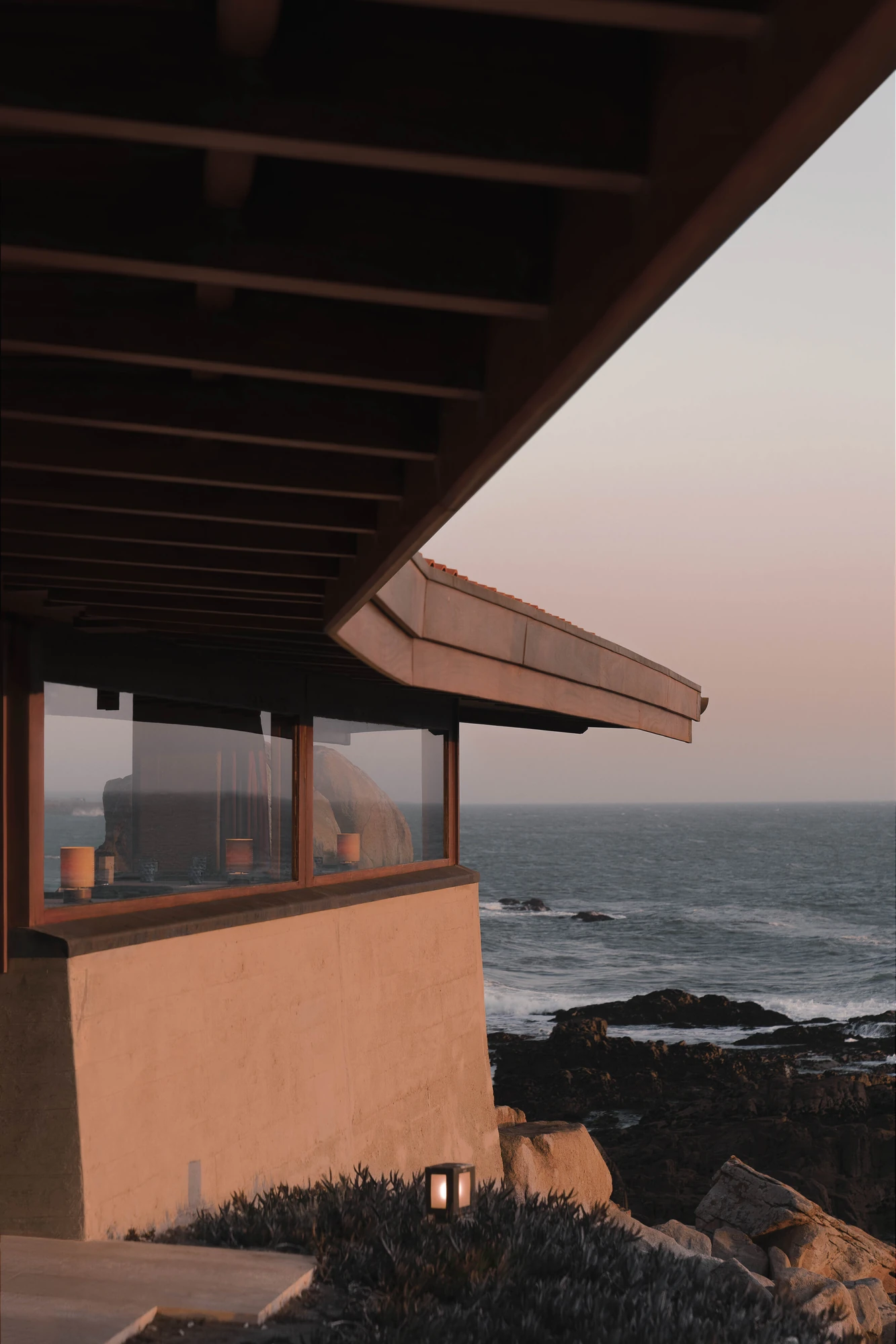 Casa de Chá da Boa Nova by Álvaro Siza, photographed by Nick Dearden — The underside of the timber roof, its rafters and pale soffit cantilevering out over a band of glazing and a rendered wall, with rocks and open sea at sunset beyond.