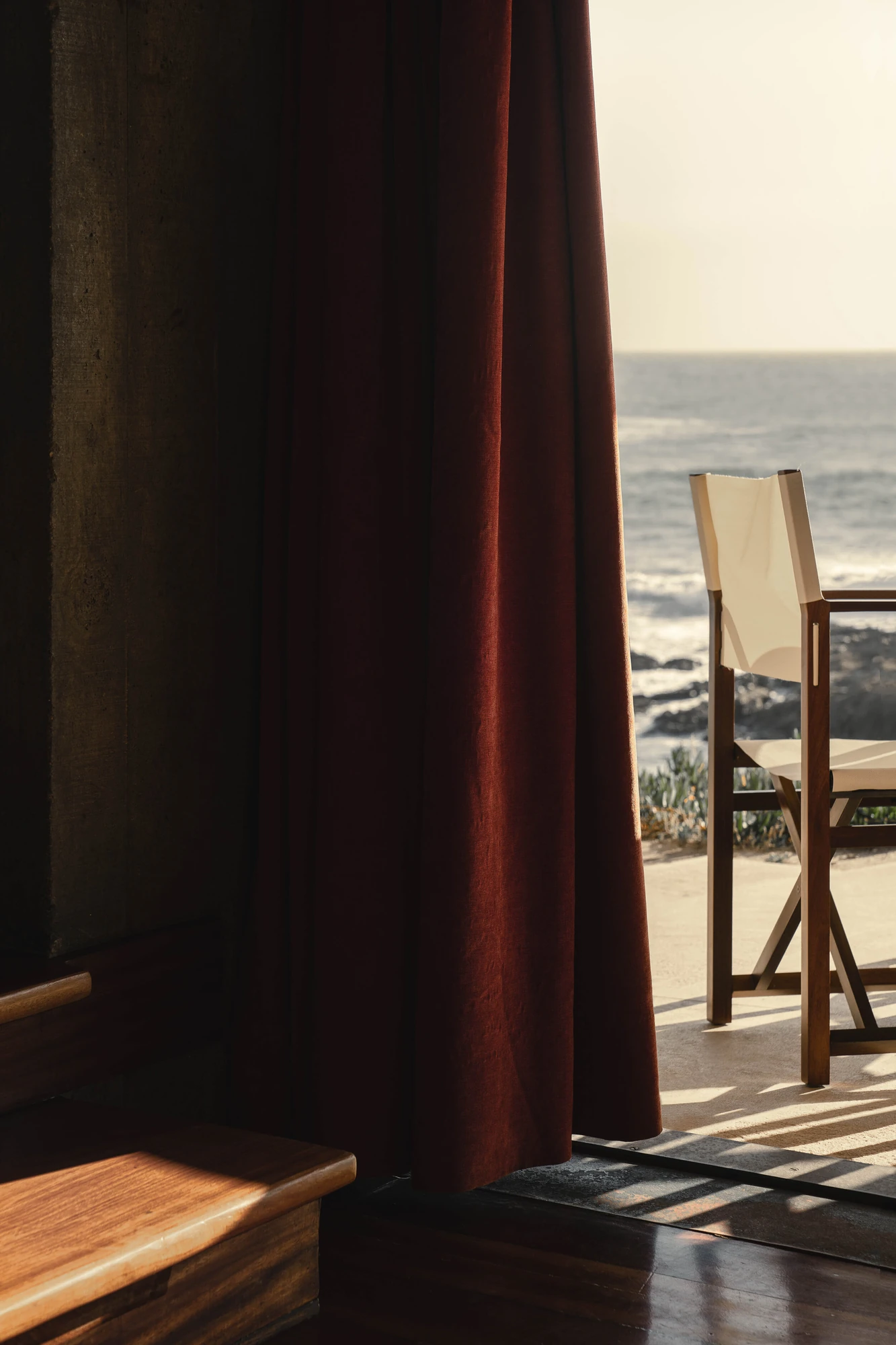 Casa de Chá da Boa Nova by Álvaro Siza, photographed by Nick Dearden — A heavy wine-red curtain hanging against a board-marked concrete wall, with low evening light falling across a timber step and a canvas and wood folding chair on the terrace.