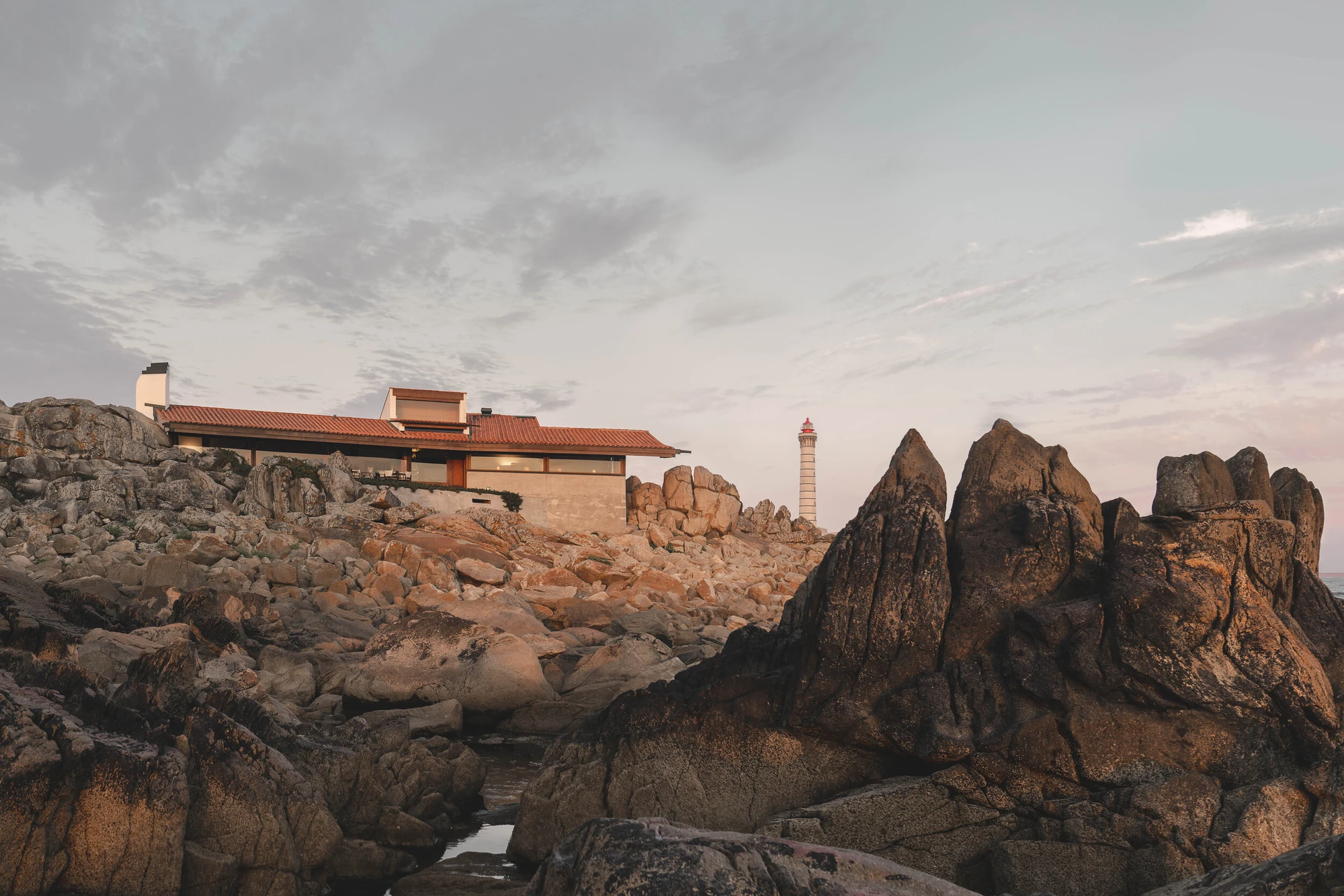 Casa de Chá da Boa Nova by Álvaro Siza, photographed by Nick Dearden — The tea house seen across a field of granite at dusk, its lit glazed dining room under a long red tile roof, with a striped lighthouse standing on the shore beyond.