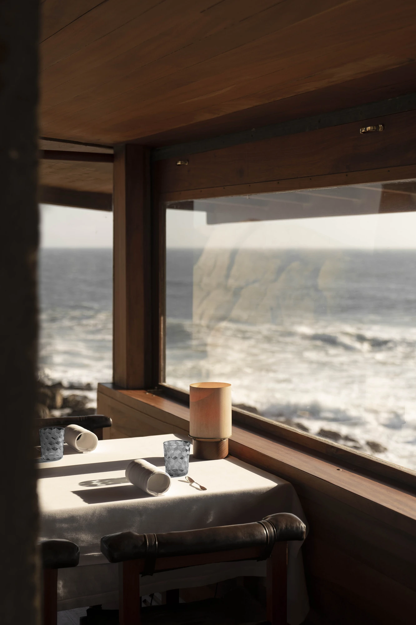 Casa de Chá da Boa Nova by Álvaro Siza, photographed by Nick Dearden — A corner table in white linen with faceted glass tumblers and a cylindrical lamp, set against a timber-framed window and the breaking Atlantic beyond.