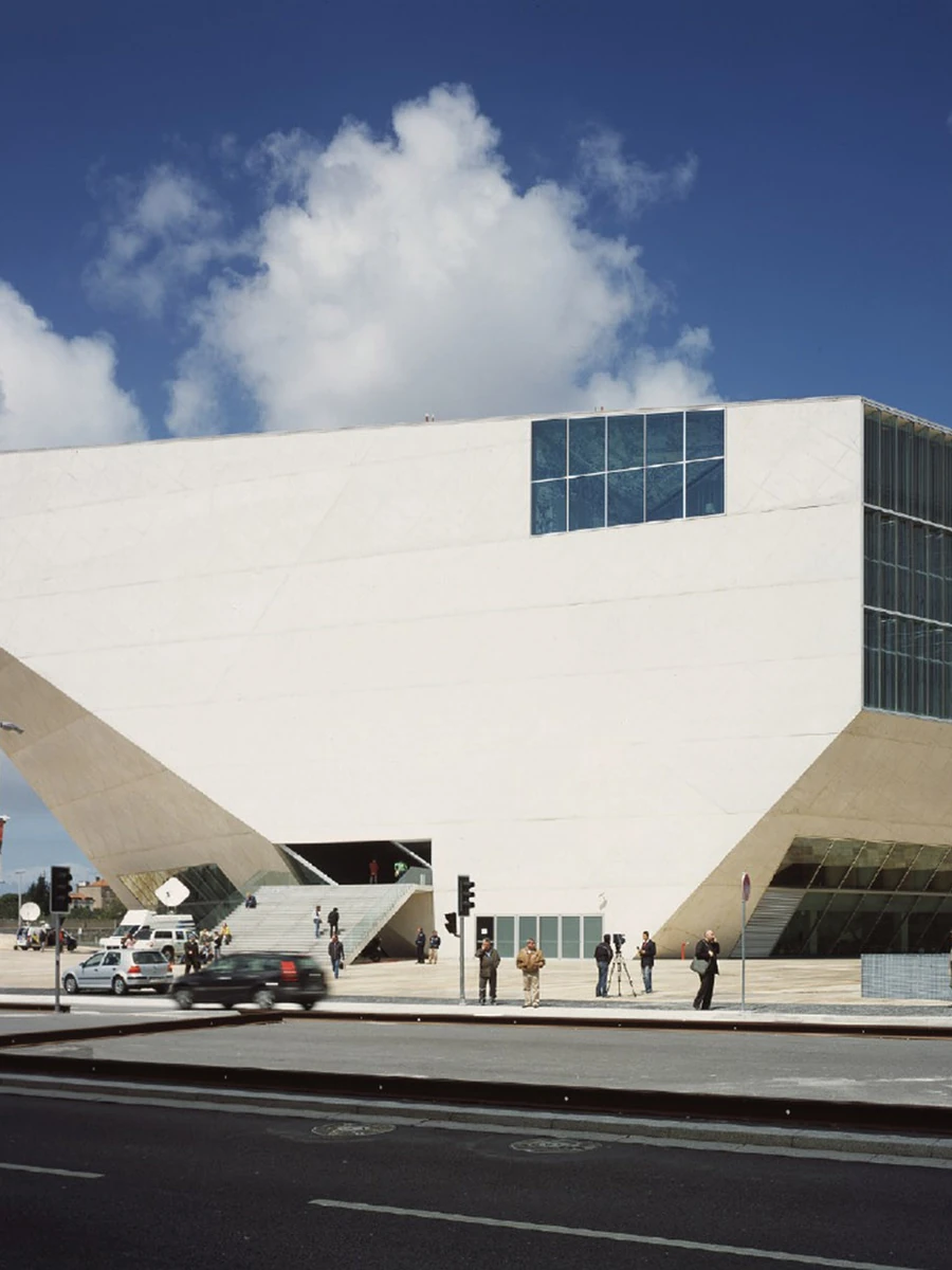 Casa da Música by OMA, photographed by Philippe Ruault — Casa da Música at dusk, its faceted white concrete mass rising above a curved canopy of shopfronts, the corrugated glass wall of the main hall lit from within.