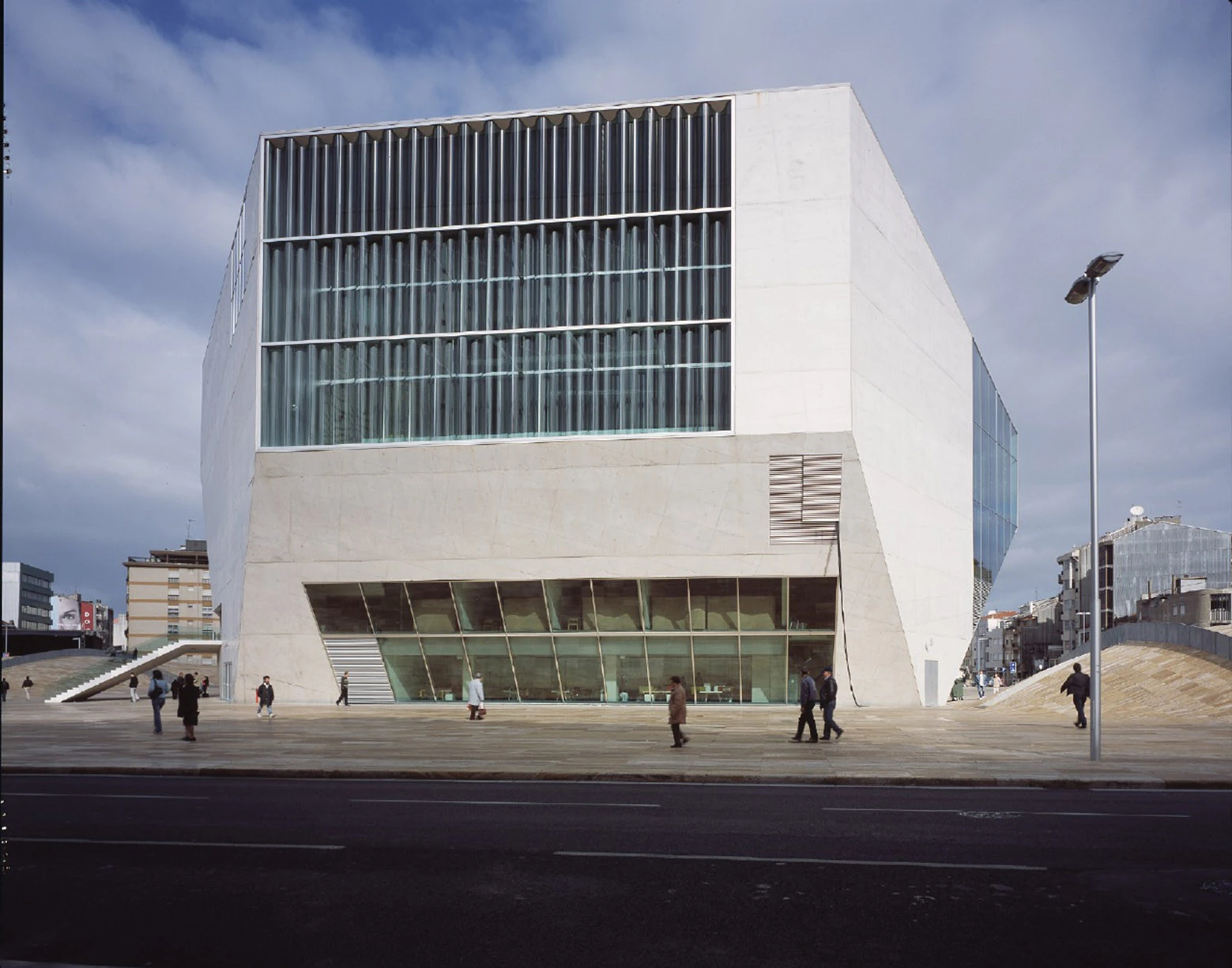 Casa da Música by OMA, photographed by Philippe Ruault — Frontal view across the travertine plaza, the corrugated glass facade of the main hall filling the upper half of the block and pedestrians crossing below.