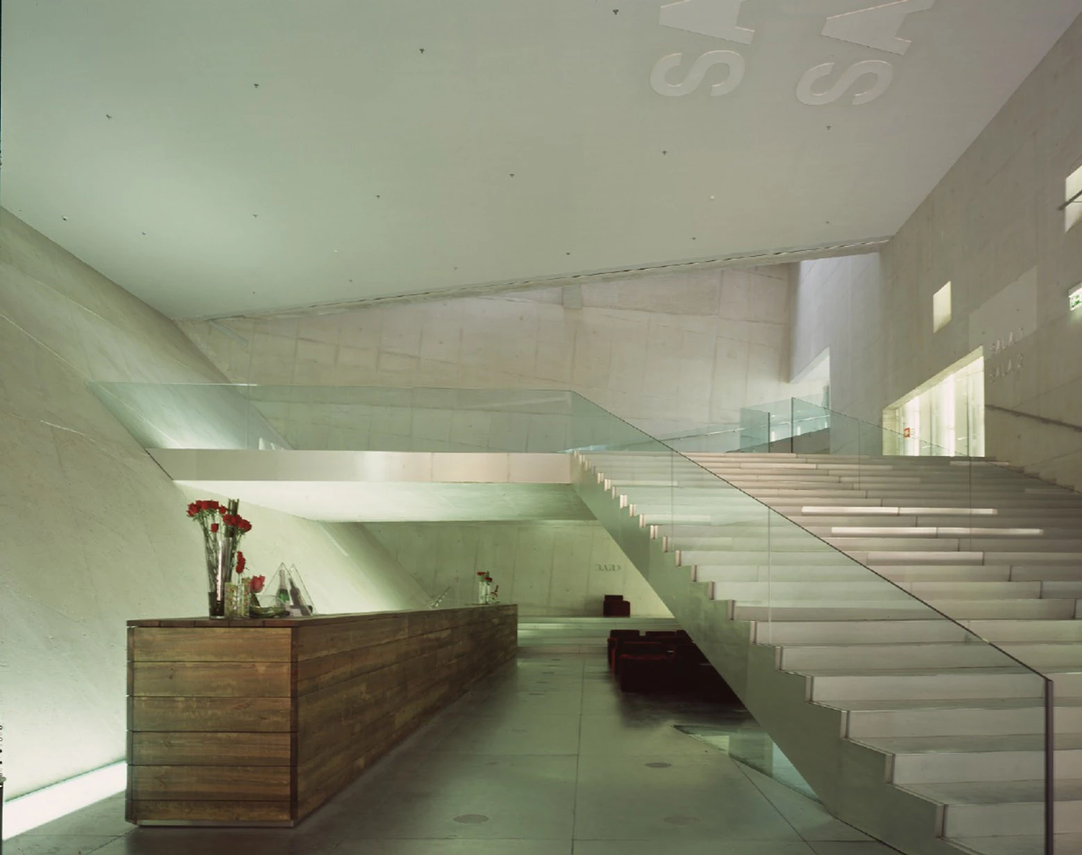 Casa da Música by OMA, photographed by Philippe Ruault — Board formed concrete foyer with a white stair and glass balustrade, a timber bar counter with red roses in the foreground and lettering set into the ceiling.
