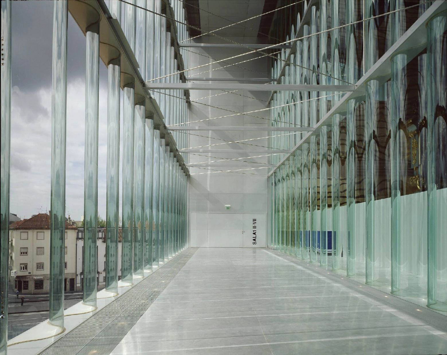 Casa da Música by OMA, photographed by Philippe Ruault — Corridor lined on both sides with vertical glass tubes, the tiled facades of Porto visible to the left and the gold grained wall of the main hall showing through to the right.
