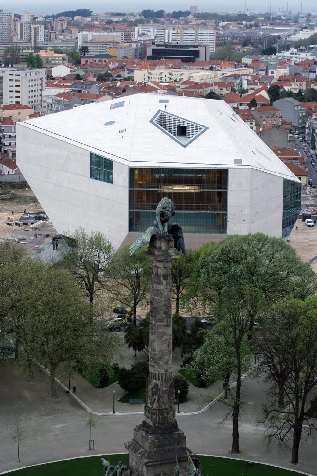 Casa da Música by OMA, photographed by Philippe Ruault — Aerial view over the Boavista park, the bronze lion on its column in the foreground and the trapezoid terrace cut into the white roof plane beyond.