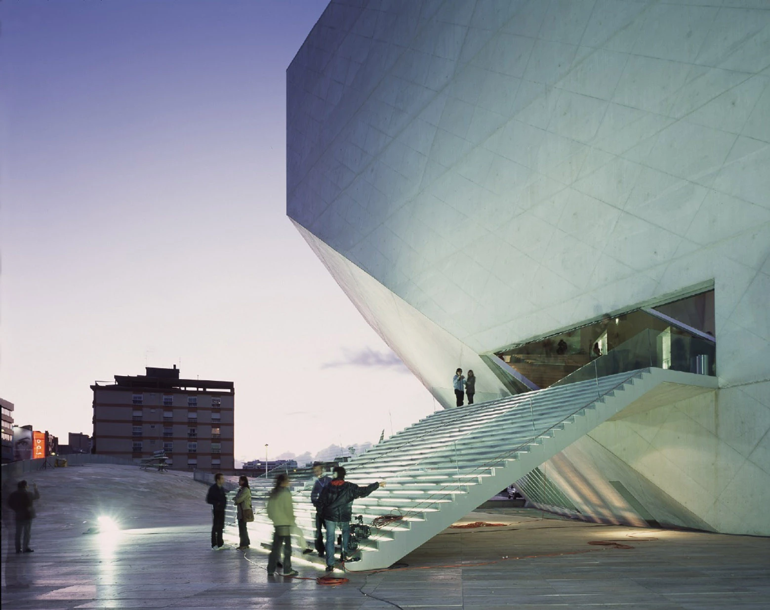 Casa da Música by OMA, photographed by Philippe Ruault — A glass balustraded stair climbing into the underside of the building at dusk, people gathered on the travertine at its foot.