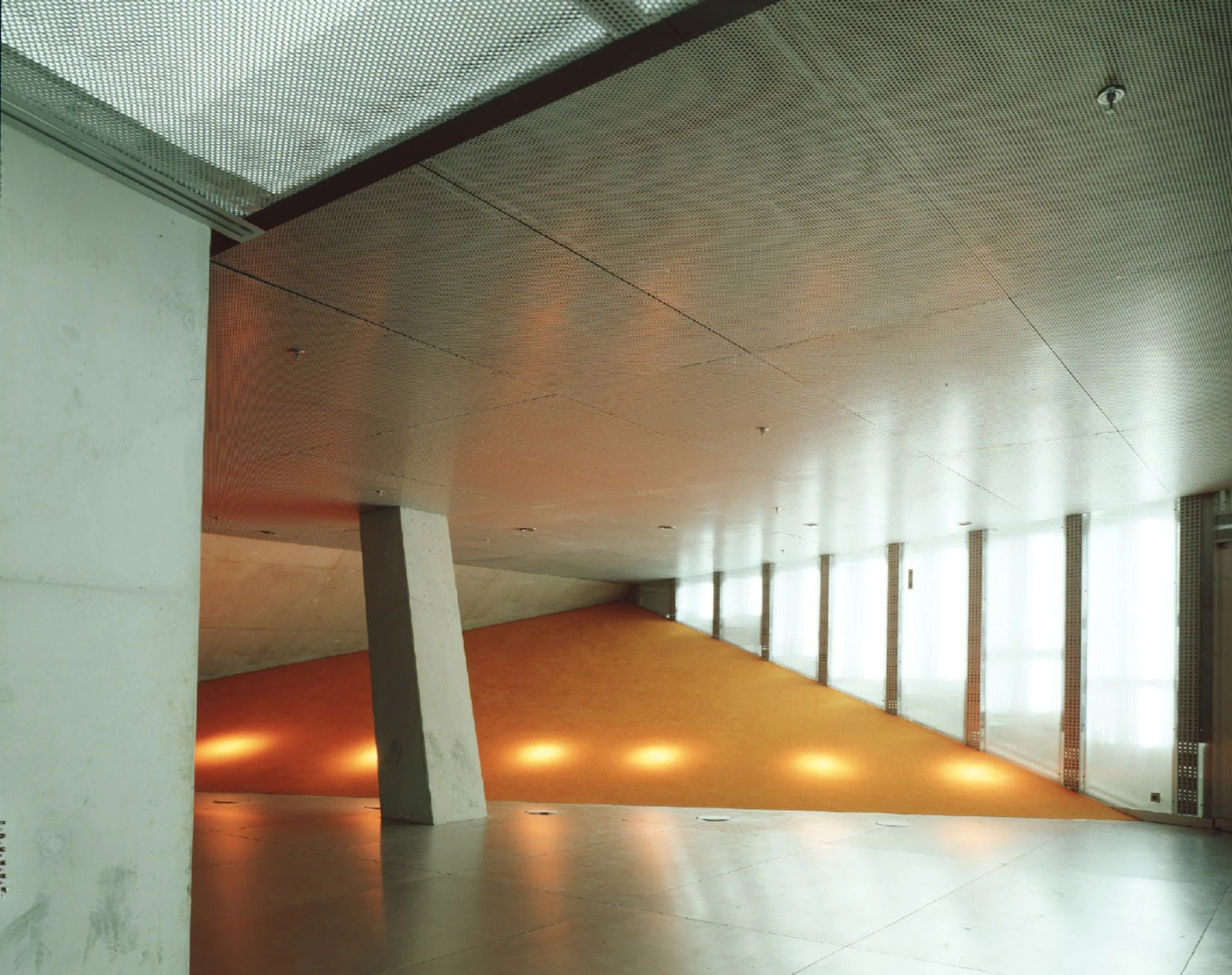 Casa da Música by OMA, photographed by Philippe Ruault — A landing where an orange floor runs under a perforated metal ceiling, a tilted concrete column at the centre and translucent glazing along the far wall.