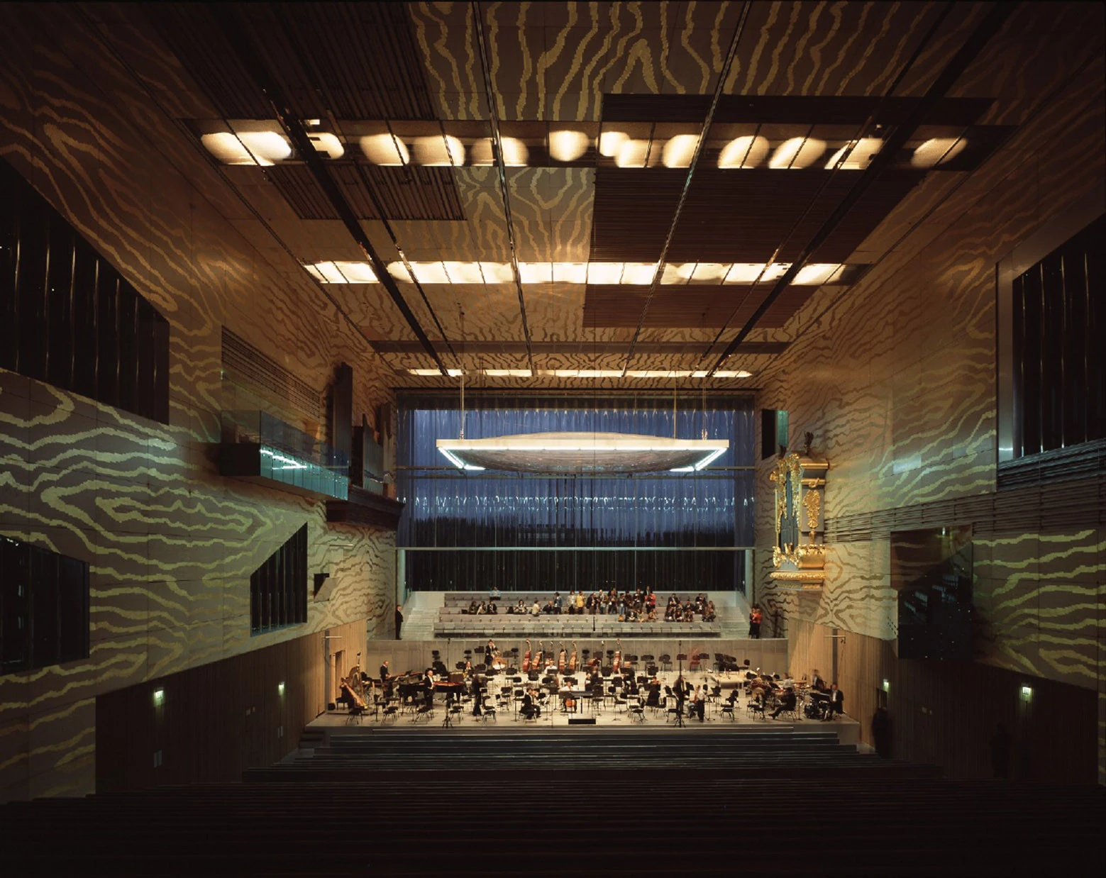 Casa da Música by OMA, photographed by Philippe Ruault — The Grand Auditorium during a performance, gold embossed plywood on every wall, corrugated glass at the far end and a flat white reflector suspended above the stage.