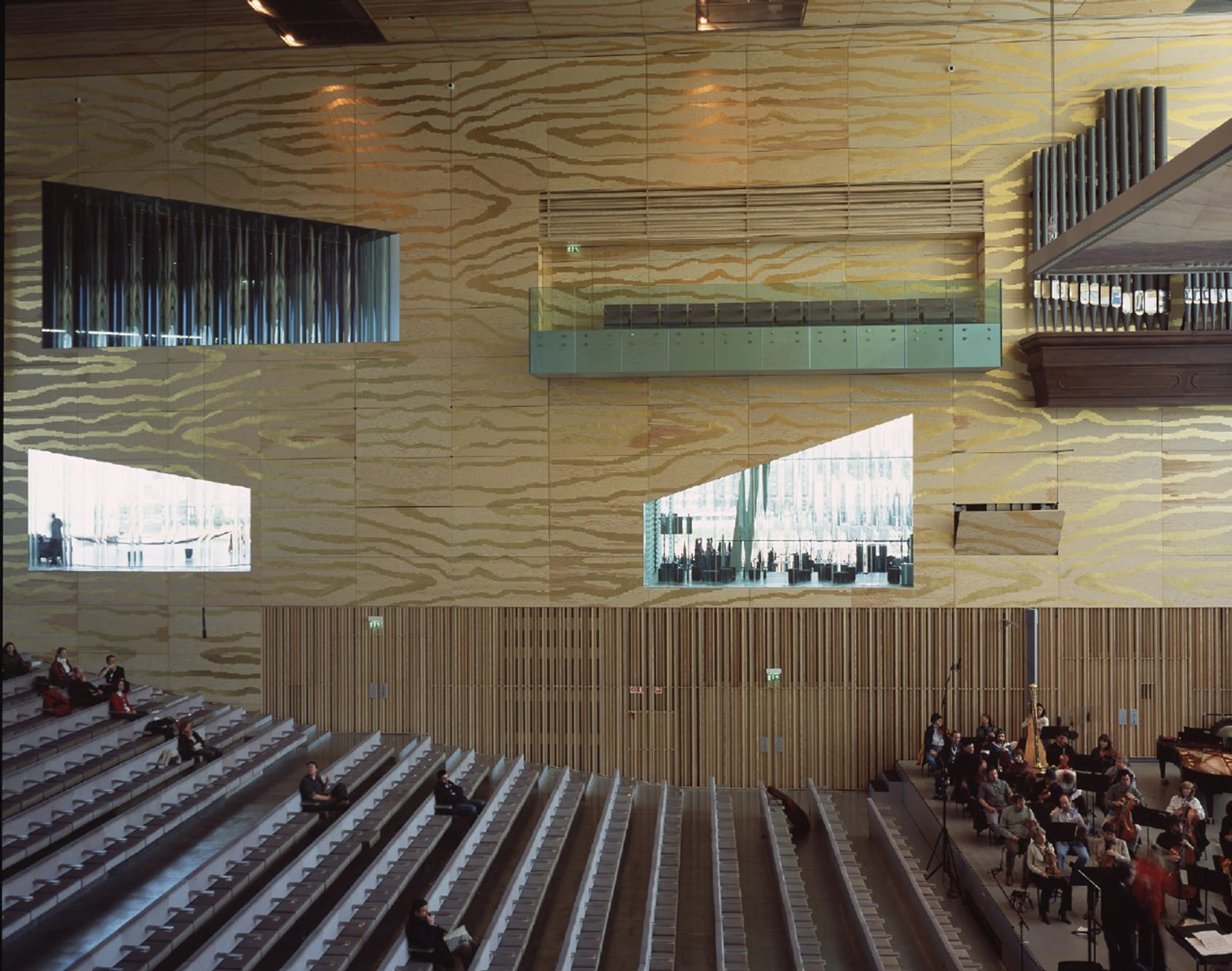 Casa da Música by OMA, photographed by Philippe Ruault — Raked seating and the gold grained plywood wall of the main hall, punctured by glazed openings onto foyers and lit rooms beyond, organ pipes at the right.