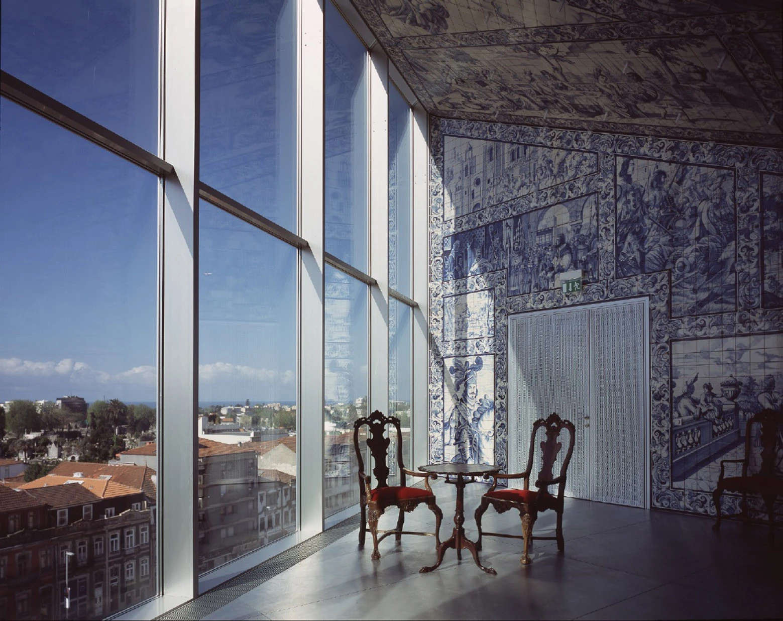 Casa da Música by OMA, photographed by Philippe Ruault — A room lined floor to ceiling in blue and white hand painted tiles, carved wooden chairs and a tripod table at a full height glazed corner over Porto's rooftops.