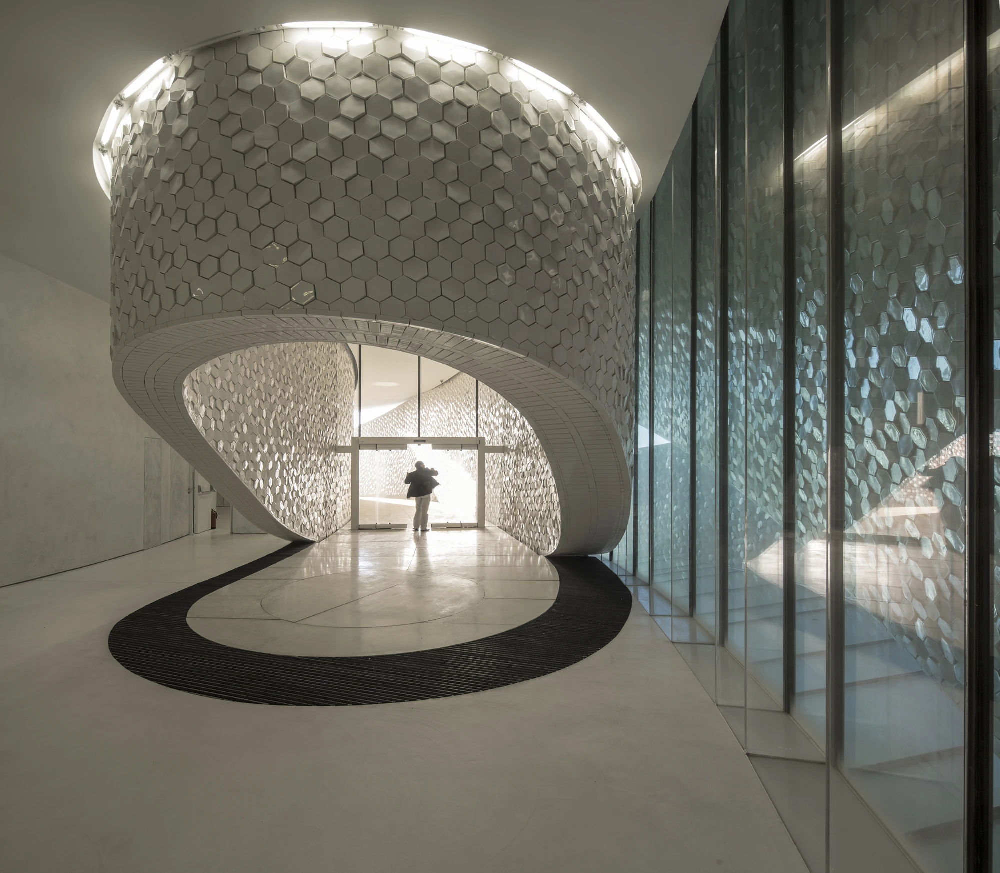 Leixões Cruise Terminal by Luís Pedro Silva Arquitecto, photographed by Fernando Guerra FG+SG — White hexagonal tiles wrap the underside of a coiling drum in the terminal lobby, a single figure silhouetted at the glazed entrance beyond and a black mat…