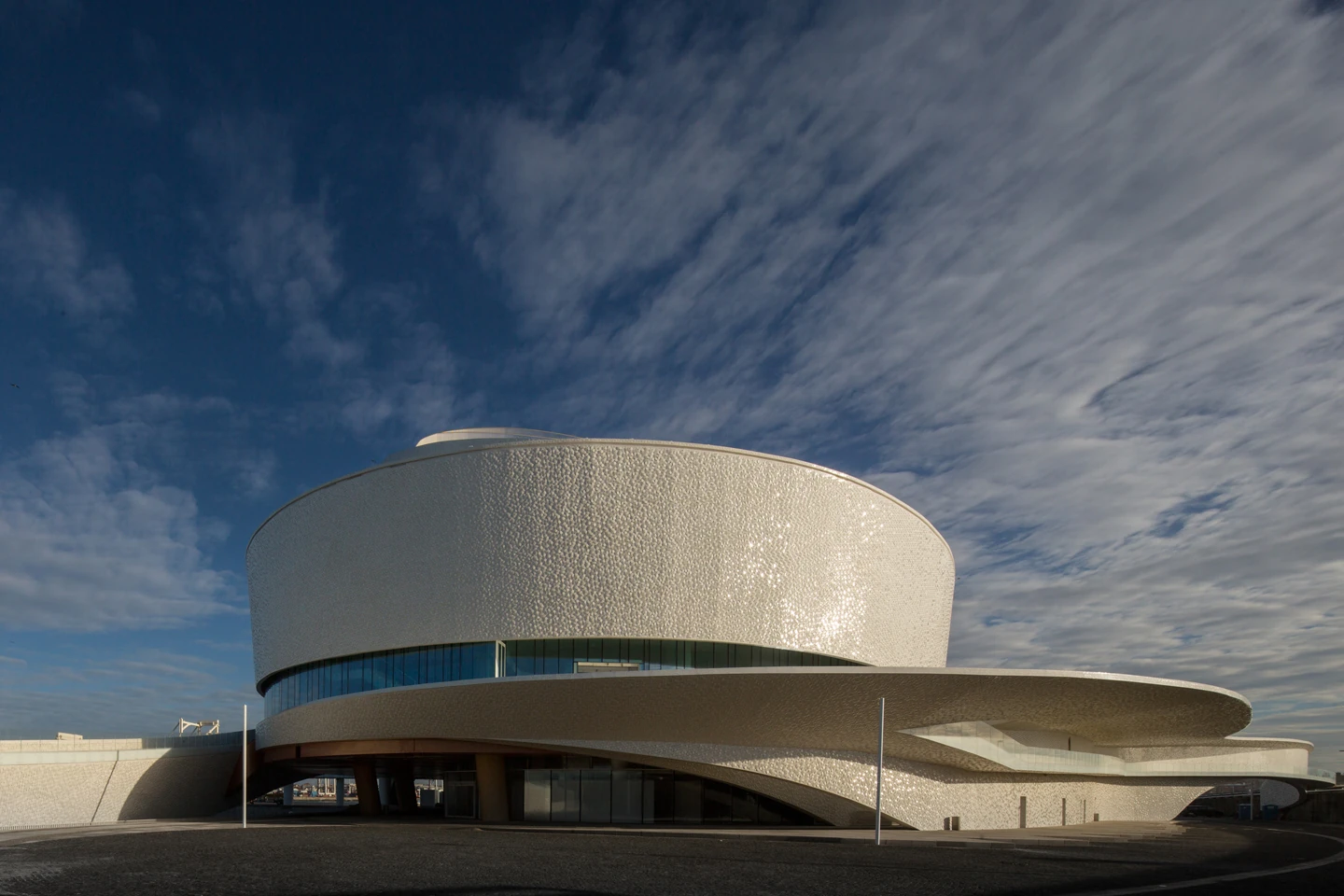 Leixões Cruise Terminal by Luís Pedro Silva Arquitecto, photographed by Fernando Guerra FG+SG — Late sun across the tiled drum of the terminal, a horizontal band of glazing beneath it and the ramp sweeping away to the right under a streaked sky.