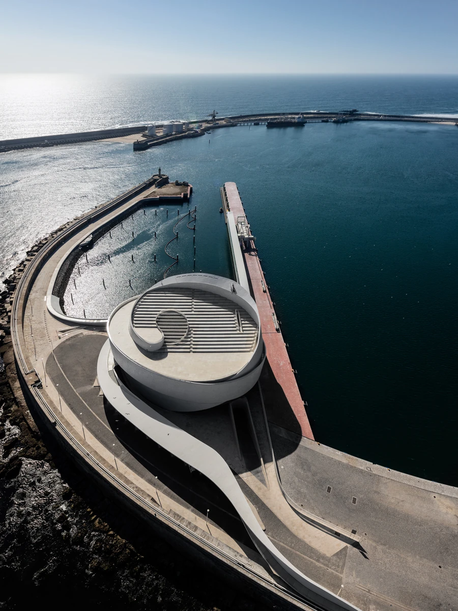 Leixões Cruise Terminal by Luís Pedro Silva Arquitecto, photographed by Fernando Guerra FG+SG — Seen from above, the white coiled terminal sits at the end of the south jetty at Leixões, its stepped roof and looping ramp turning between the marina basin…