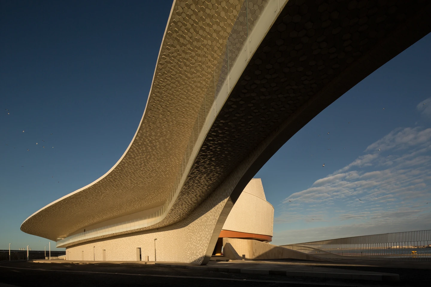 Leixões Cruise Terminal by Luís Pedro Silva Arquitecto, photographed by Fernando Guerra FG+SG — The elevated ramp sweeps overhead, its hexagon-tiled soffit turning gold in low sun above the tiled base of the drum.