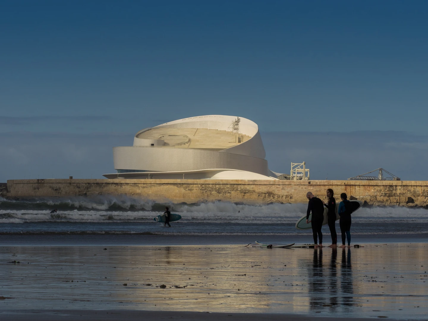 Leixões Cruise Terminal by Luís Pedro Silva Arquitecto, photographed by Fernando Guerra FG+SG — The white mass of the terminal rises behind the granite breakwater while surfers stand on the wet sand at Matosinhos beach.