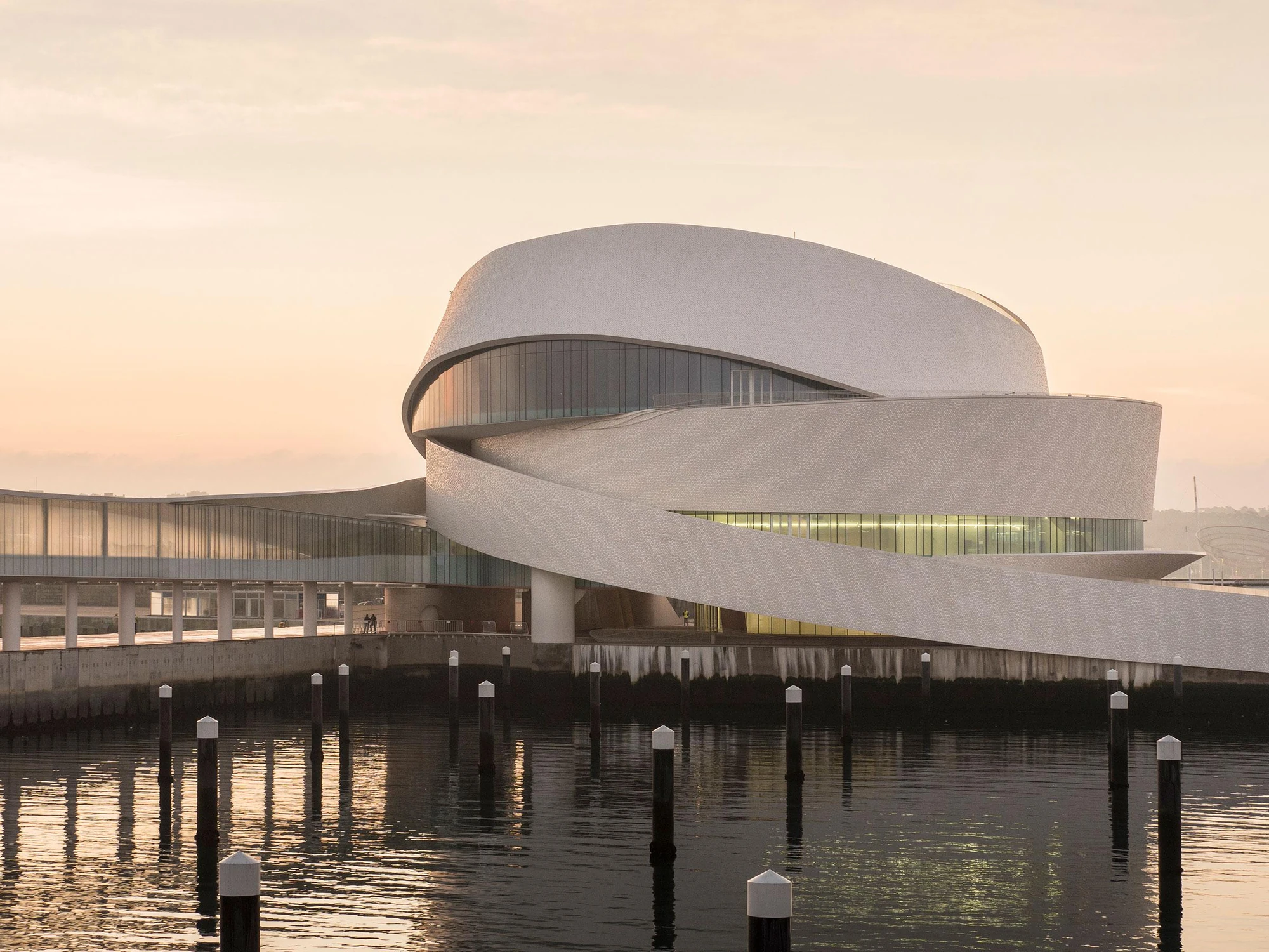 Leixões Cruise Terminal by Luís Pedro Silva Arquitecto, photographed by Fernando Guerra FG+SG — The terminal at dawn across the marina moorings, tiled bands spiralling above a lit band of glazing, timber piles standing in still water in the foreground.