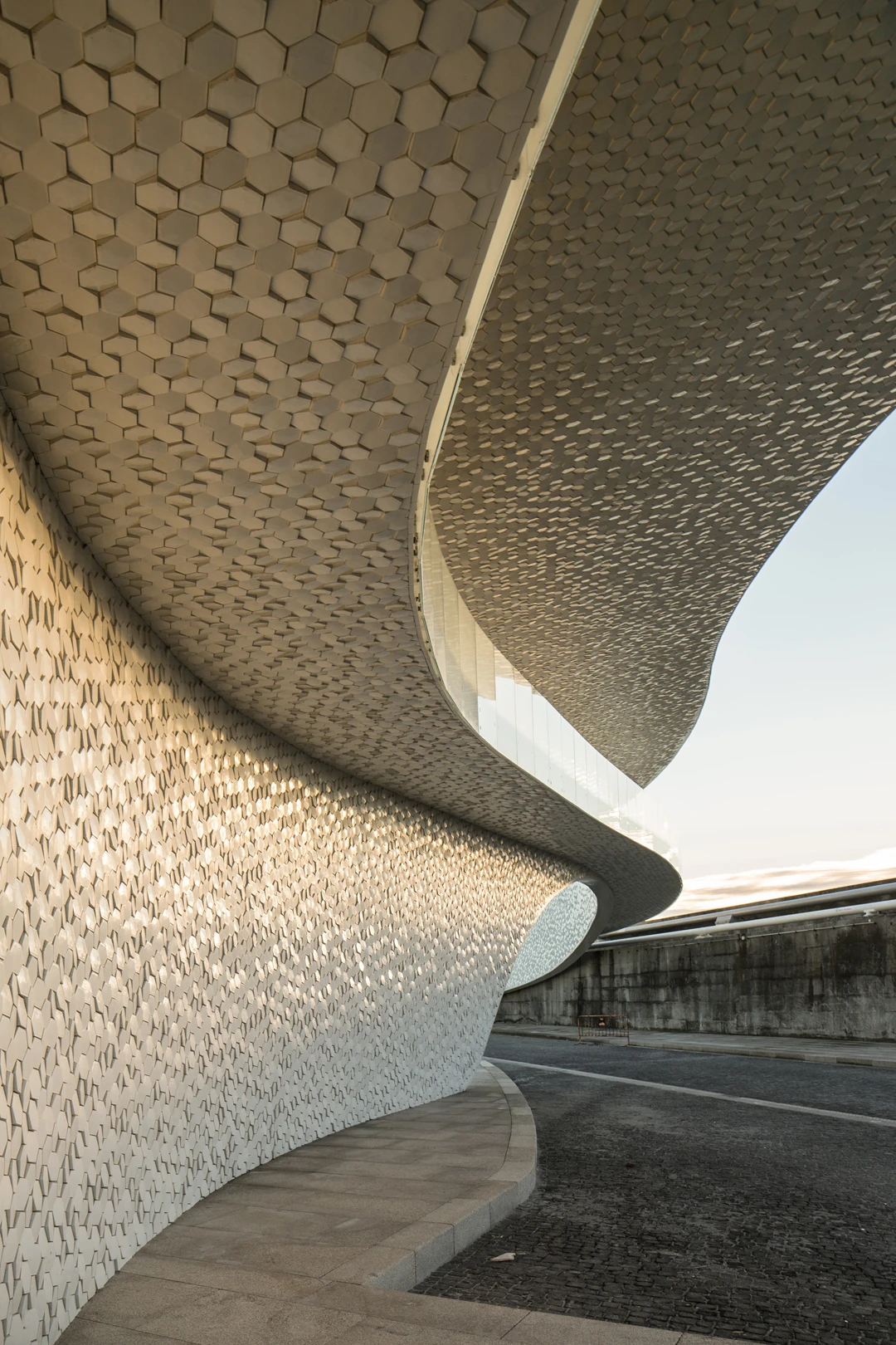 Leixões Cruise Terminal by Luís Pedro Silva Arquitecto, photographed by Fernando Guerra FG+SG — Two tiled surfaces meet where the ramp lifts away from the wall, hexagons in deep relief above and a finer scaled tile below, the road curving past at ground…
