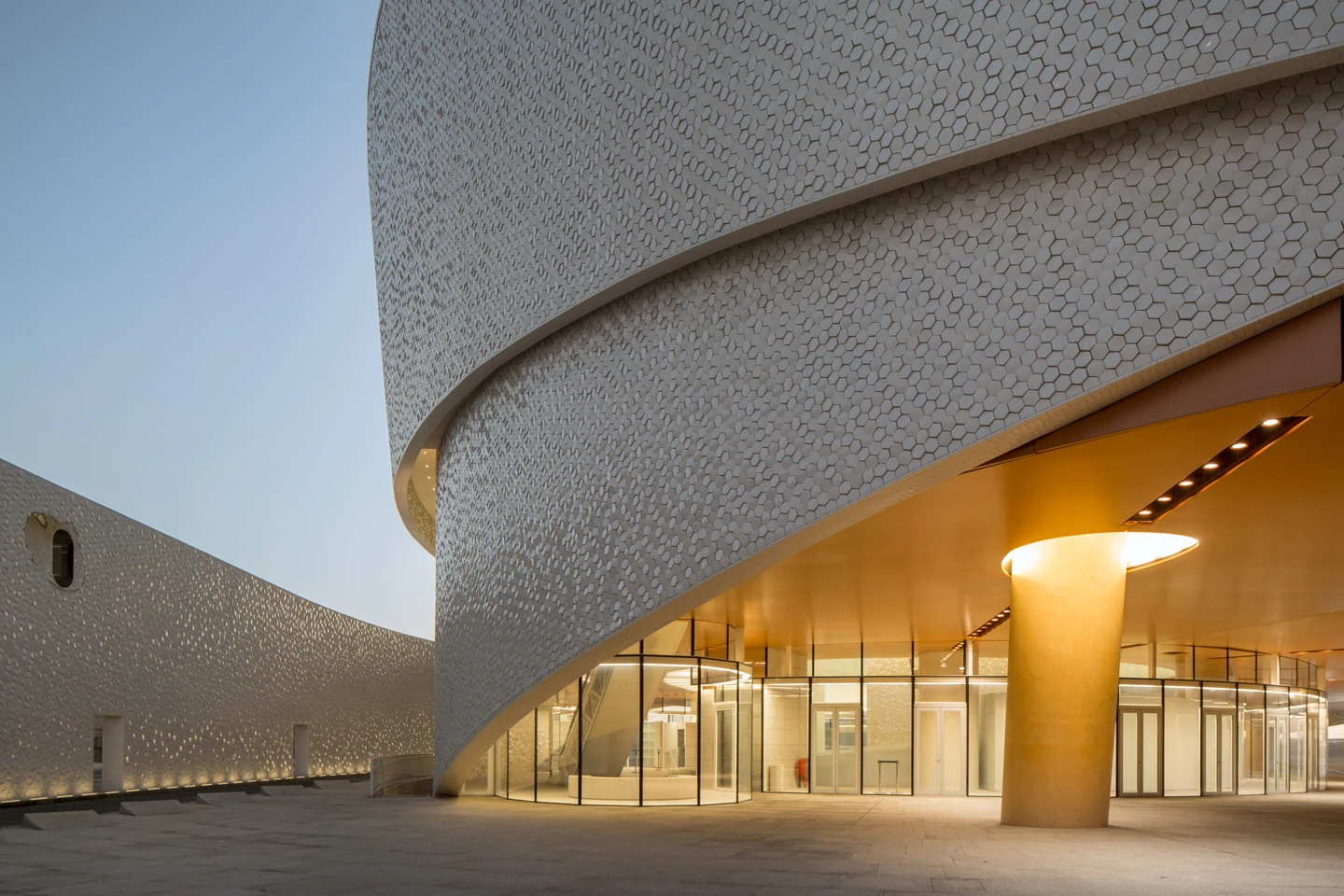 Leixões Cruise Terminal by Luís Pedro Silva Arquitecto, photographed by Fernando Guerra FG+SG — Evening at the base of the terminal, warm light spilling from the fully glazed ground floor beneath the tiled overhang and a tapered column lit from within.