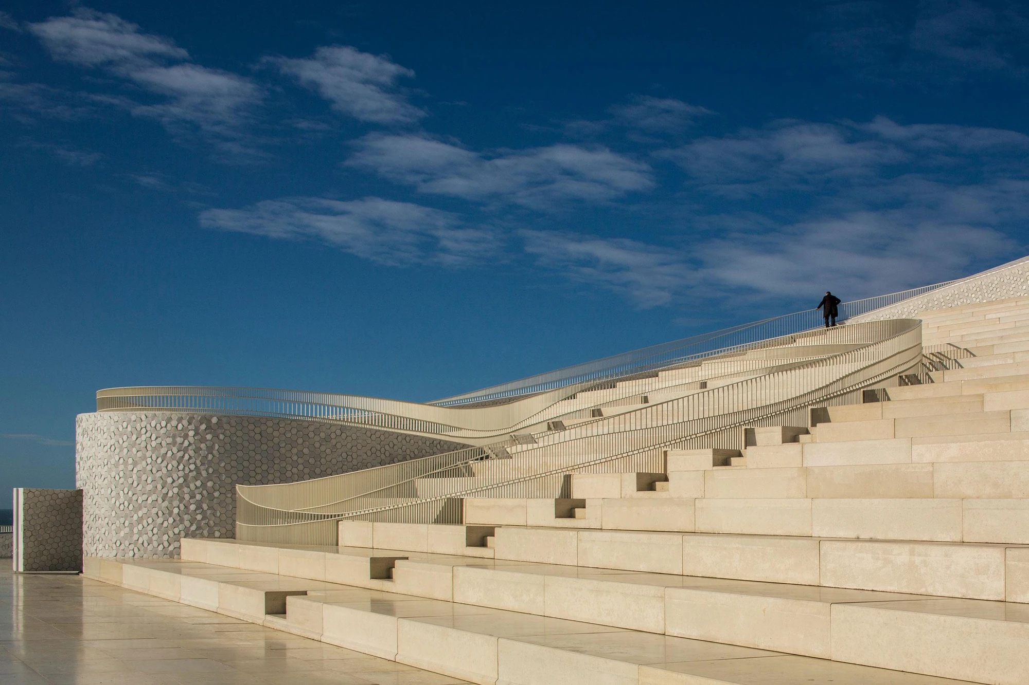 Leixões Cruise Terminal by Luís Pedro Silva Arquitecto, photographed by Fernando Guerra FG+SG — Broad steps of pale stone rise across the roof of the terminal against a deep blue sky, a ribbon of railing threading through them and one figure at the crest.