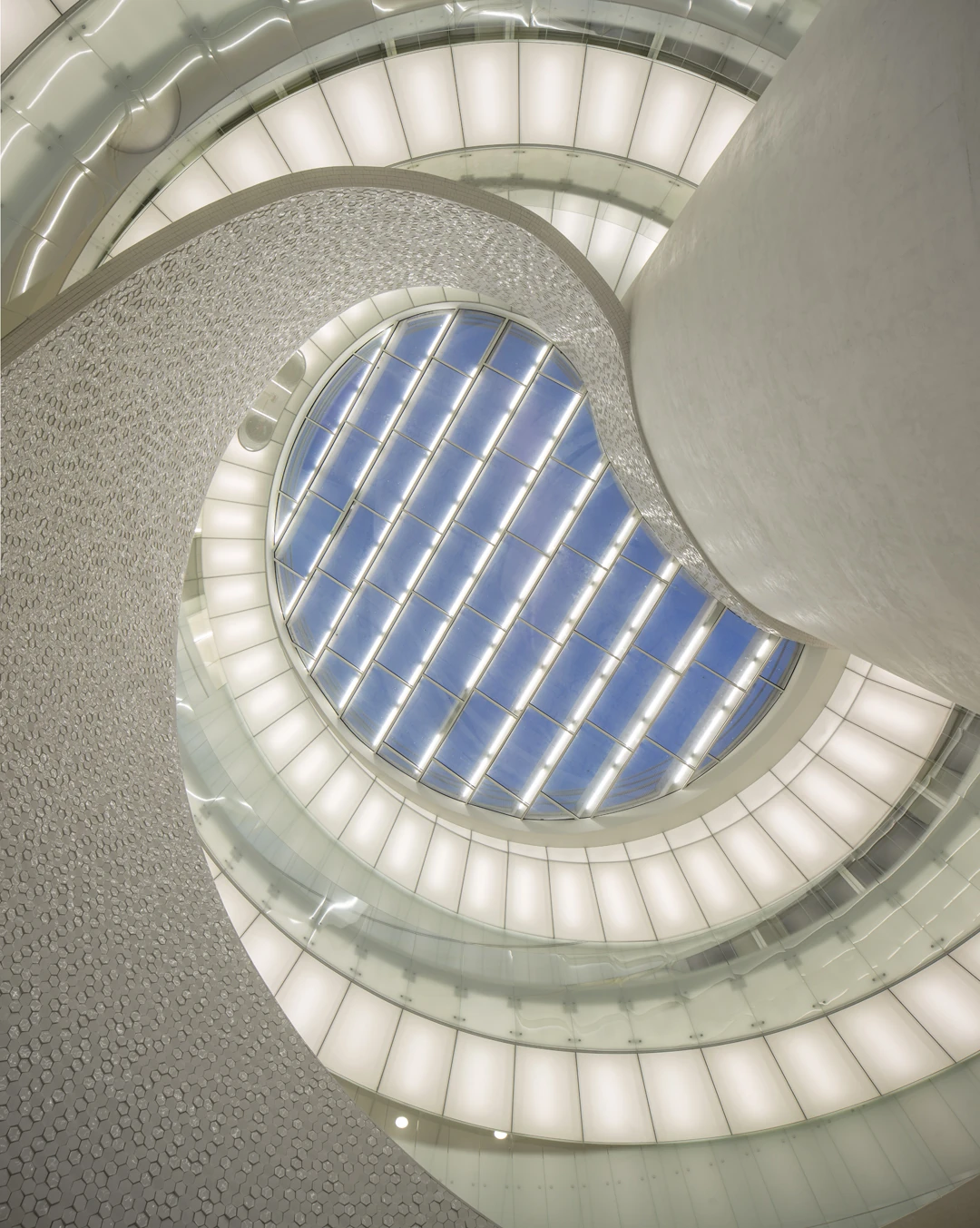 Leixões Cruise Terminal by Luís Pedro Silva Arquitecto, photographed by Fernando Guerra FG+SG — Looking up through the helical ramp to the oval skylight at the top of the quadruple-height core, tiled wall on one side and lit glass balustrades spiralling…