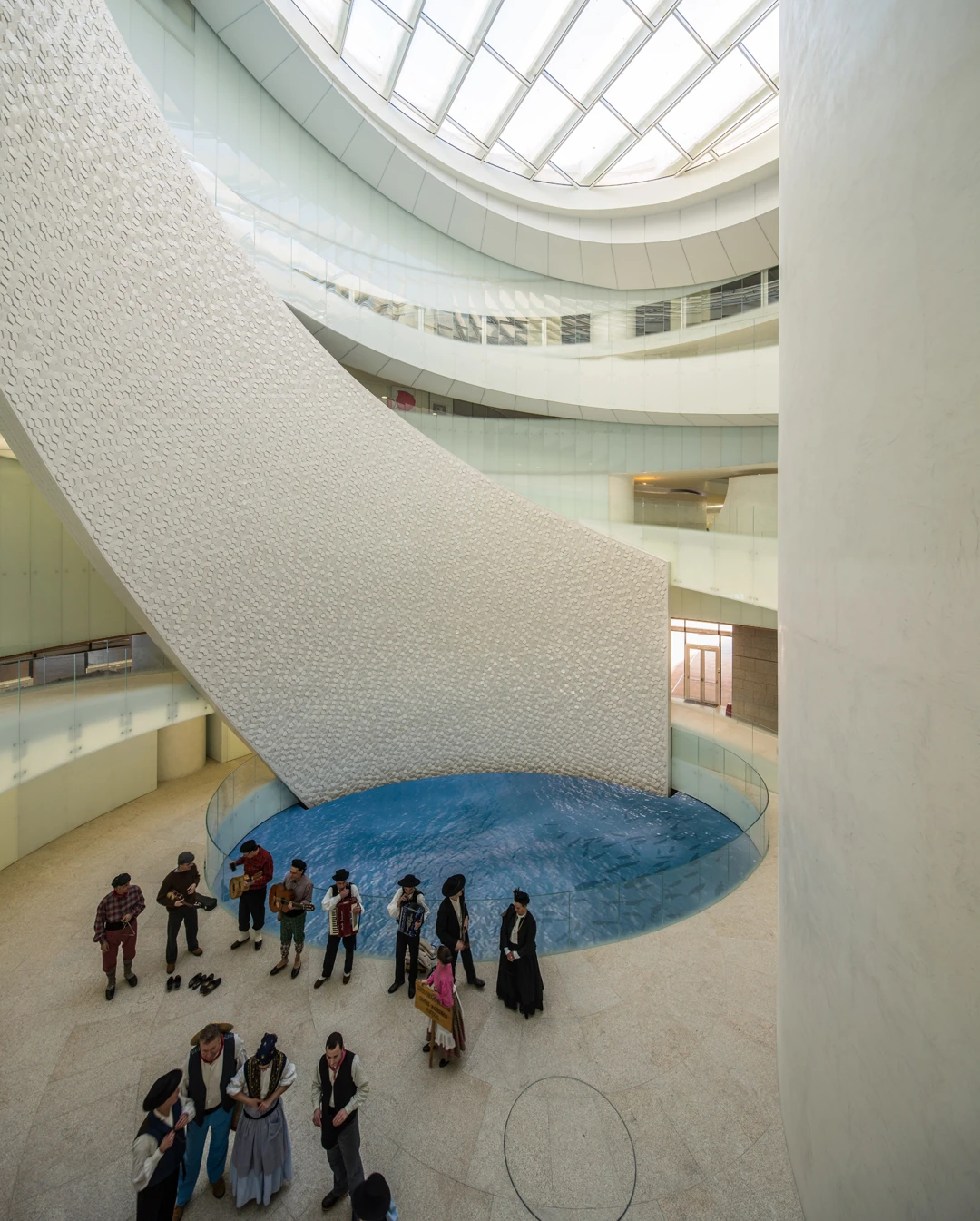 Leixões Cruise Terminal by Luís Pedro Silva Arquitecto, photographed by Fernando Guerra FG+SG — Musicians in traditional dress stand at the edge of the shallow blue pool at the base of the atrium, the tiled ramp sweeping down toward it under the skylight.