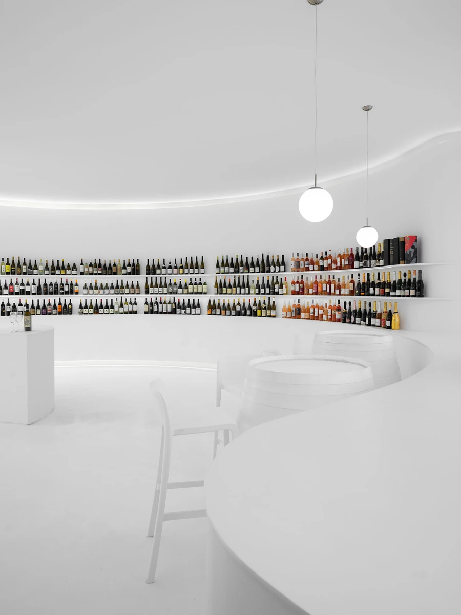 Portugal Vineyards Concept Store by Porto Architects, photographed by Ivo Tavares Studio — The curving shelves of bottles seen past a white chair and whitewashed barrel tables, a cove of concealed light running around the ceiling above.