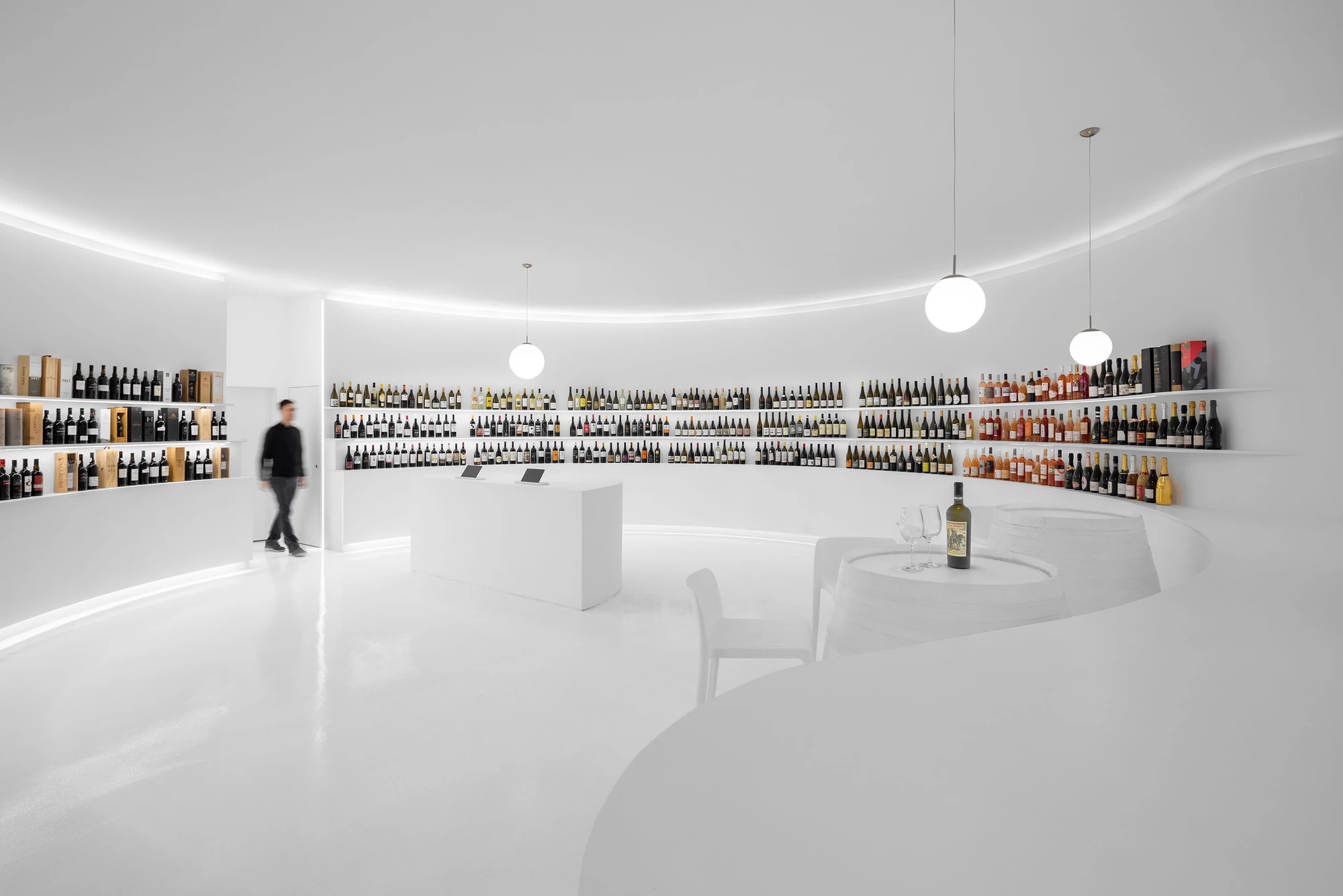 Portugal Vineyards Concept Store by Porto Architects, photographed by Ivo Tavares Studio — The full sweep of the open-plan store, a figure at a doorway on the left, the white counter at the centre and rows of bottles running along the curved wall.