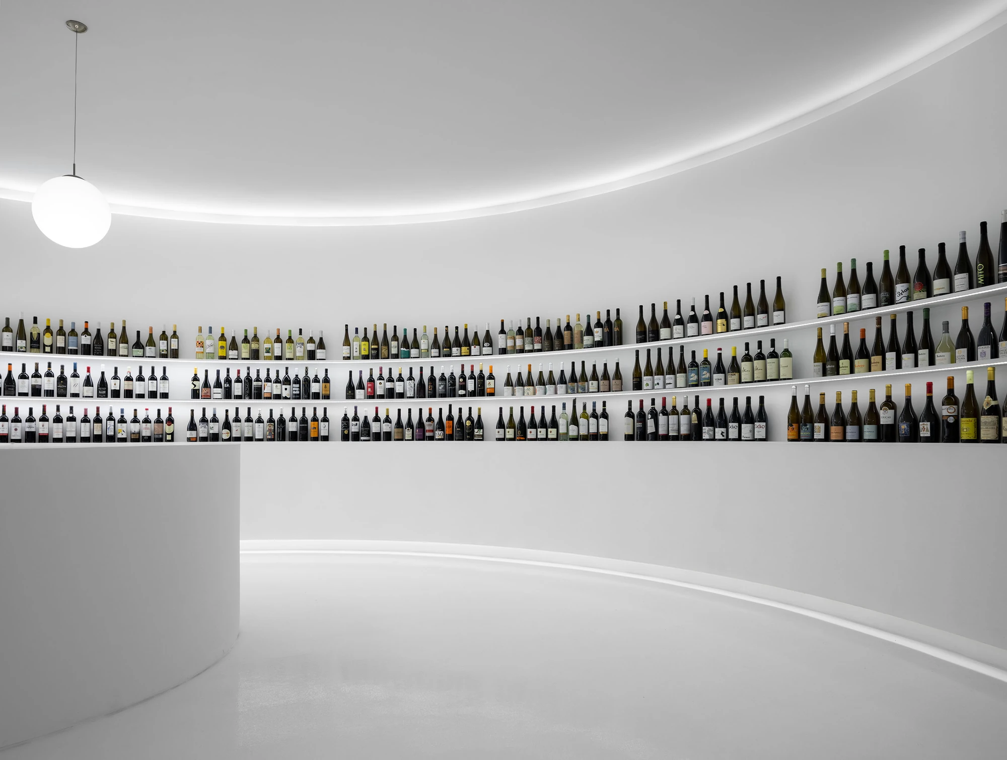 Portugal Vineyards Concept Store by Porto Architects, photographed by Ivo Tavares Studio — Three continuous shelves of Portuguese wine rise across the curved white wall, each lit from beneath, an opal glass pendant hanging at the left.