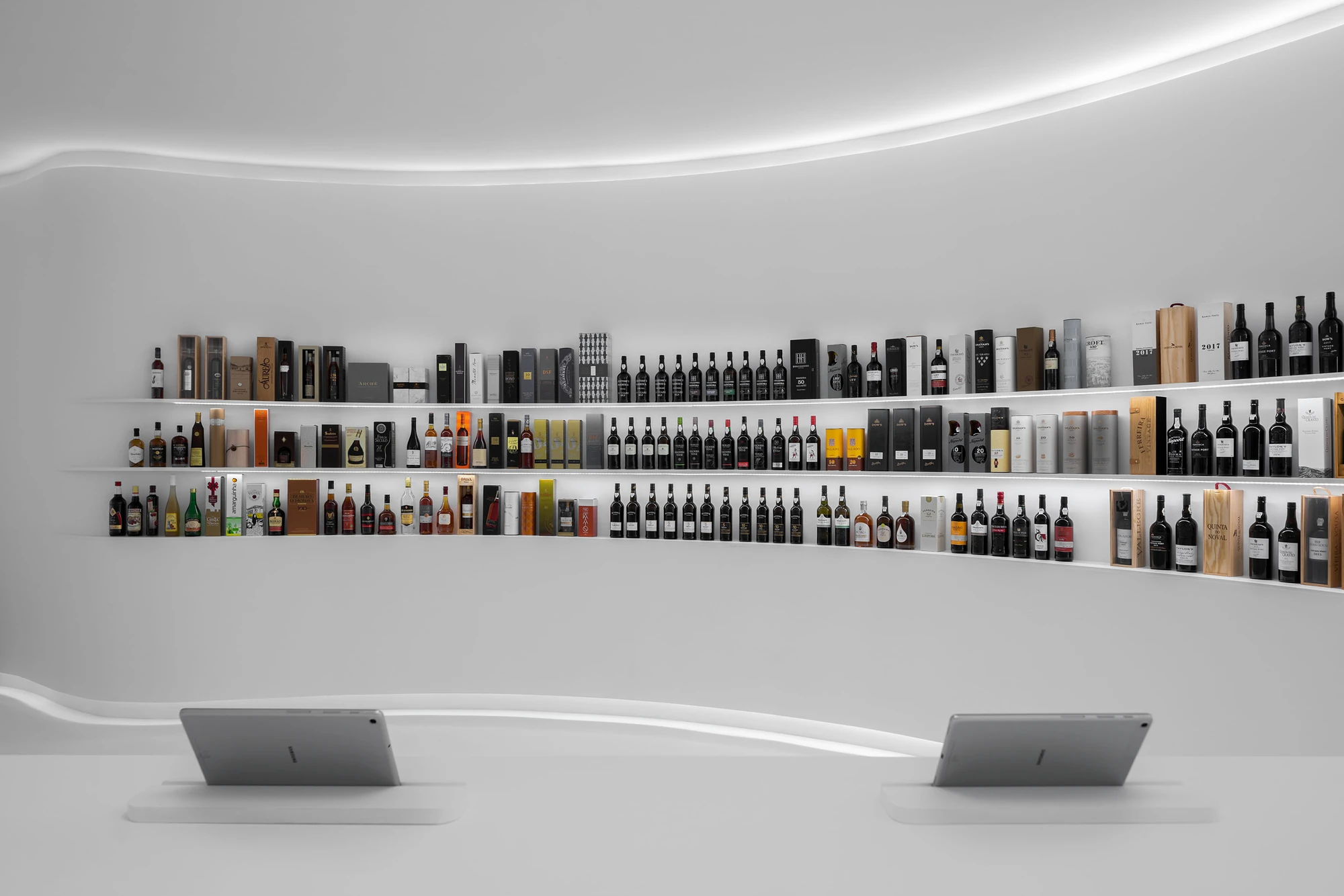 Portugal Vineyards Concept Store by Porto Architects, photographed by Ivo Tavares Studio — Two tablets sit on the white counter facing a wall of port and fortified wine in wooden presentation cases, the cove light bending above.