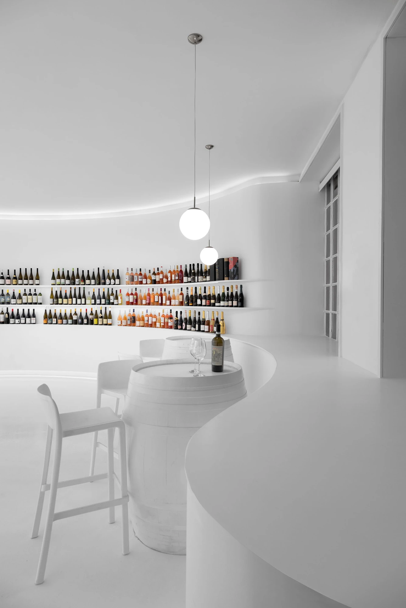 Portugal Vineyards Concept Store by Porto Architects, photographed by Ivo Tavares Studio — White stools stand at whitewashed barrel tables in the tasting area, the curved counter sweeping past and the wine wall glowing behind.