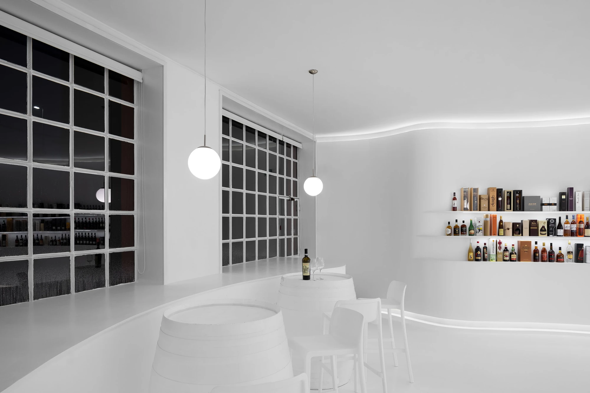 Portugal Vineyards Concept Store by Porto Architects, photographed by Ivo Tavares Studio — The tasting area at night, tall steel-framed windows black against the white room, two opal pendants hanging above painted barrels and white chairs.