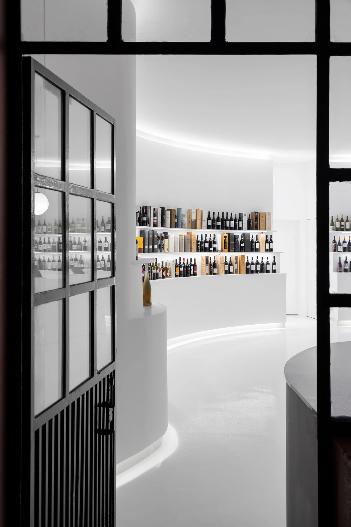 Portugal Vineyards Concept Store by Porto Architects, photographed by Ivo Tavares Studio — The store seen from outside through the open black steel door, the white curve of the counter and the lit shelves receding within.