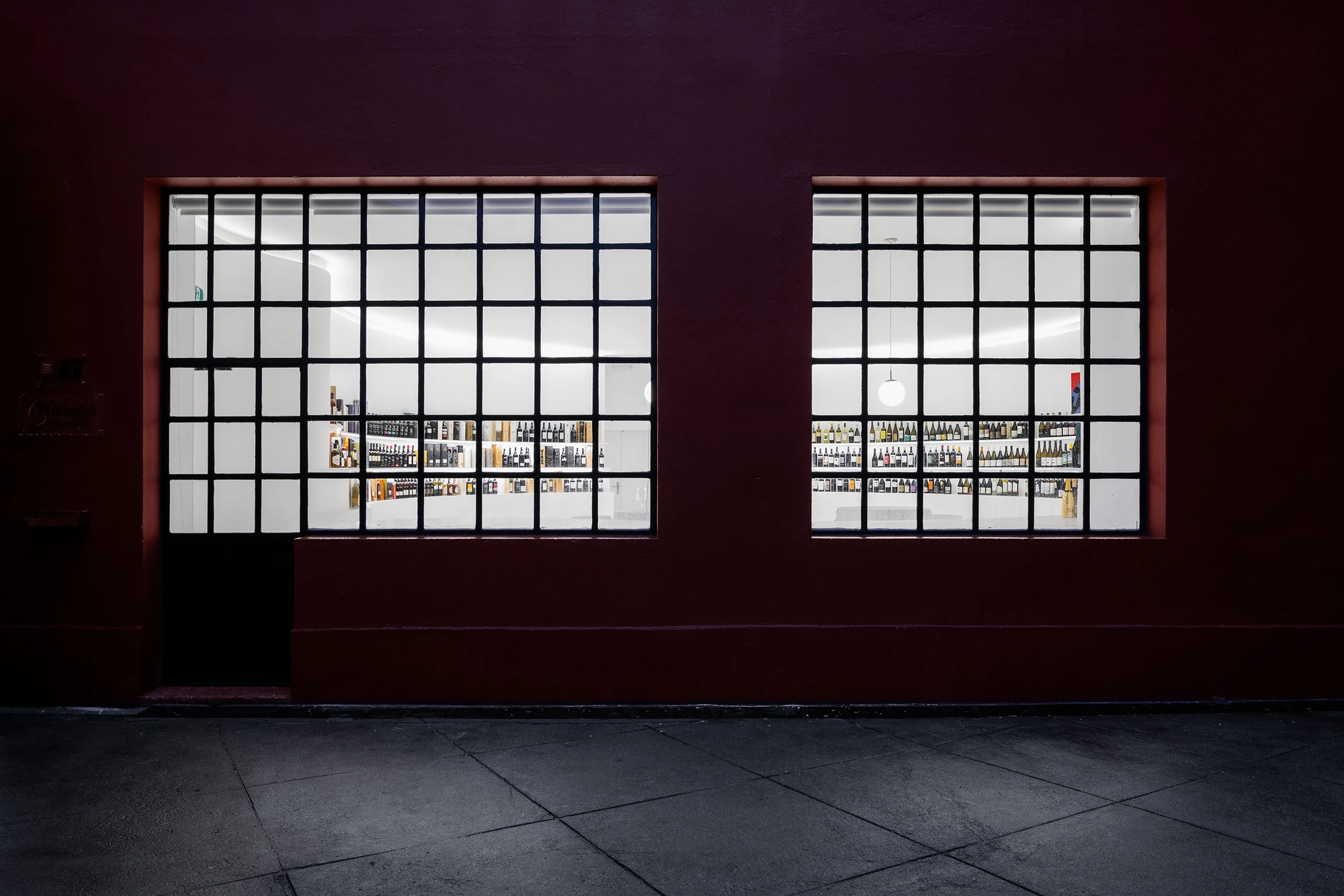 Portugal Vineyards Concept Store by Porto Architects, photographed by Ivo Tavares Studio — The oxblood street facade at night, two steel-gridded windows glowing white with rows of bottles visible inside.