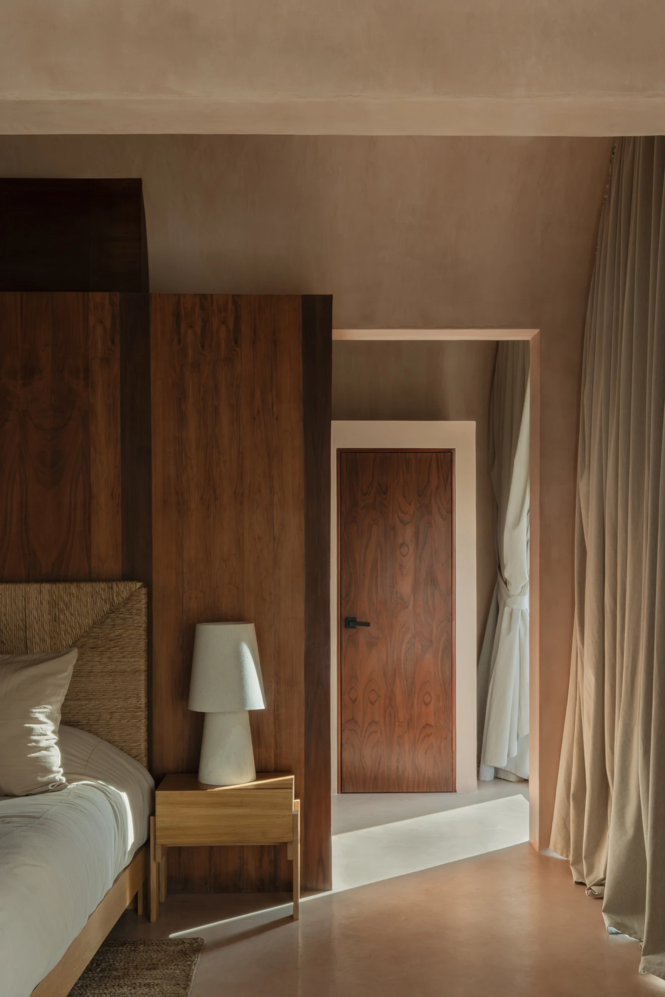 Babel by V Taller, photographed by Funciono — A bedroom with a rosewood-panelled headboard wall and matching door, woven bedhead and a ceramic lamp against plaster in a soft clay tone.